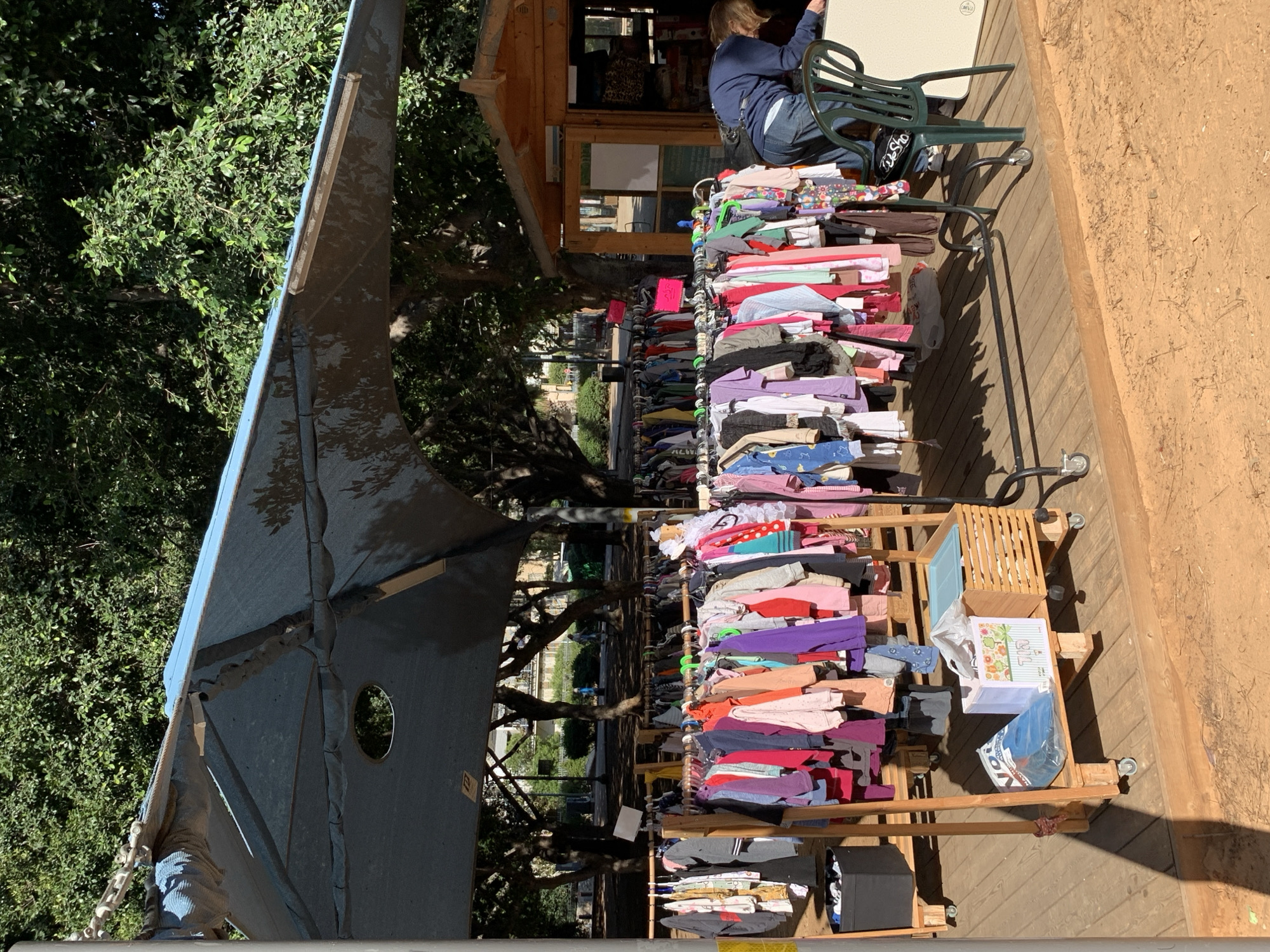 The image depicts an outdoor scene, likely a marketplace in Tel-Aviv, Israel. A woman with short blonde hair wearing a blue sweatshirt and jeans sits on a green chair next to a wooden structure. Numerous racks of clothing are displayed, filled with various colors and styles. These racks appear to be wooden, with wheels at the bottom for mobility. An awning provides shade over some of the clothing racks. Trees and greenery are visible in the background, indicating an outdoor setting. A wooden platform or sidewalk is on the right side of the image.