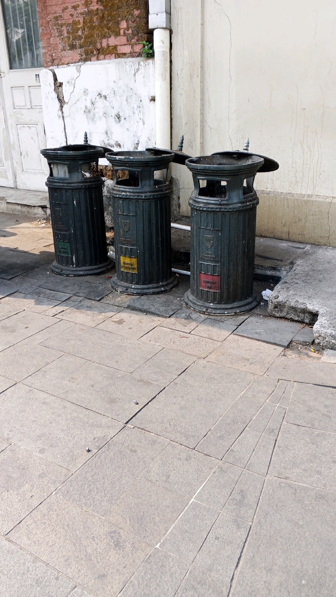 The image shows three dark green, ornate trash cans lined up on a stone sidewalk in front of a weathered brick and stucco building. The trash cans have ribbed bases and decorative tops with pointed finials. Each trash can has a label attached. The leftmost trash can has a green label. The middle trash can has a yellow label. The rightmost trash can has a red label. The building behind the trash cans is partially visible, with a white door or window on the left and a white wall with vertical streaks of discoloration on the right. A downspout runs vertically down the wall. The sidewalk is paved with large, grey, rectangular stones arranged in a geometric pattern. The scene is set outdoors in what appears to be a public urban space. The trash cans are official-looking, with crests and text that are partially visible, suggesting they are part of municipal waste management in Semarang, Indonesia. The red label on the rightmost trash can appears to have "SAMPAH" printed on it, which is Indonesian for "trash" or "garbage". The environment appears somewhat aged, with peeling paint or stucco on the building and some debris on the ground around the trash cans.