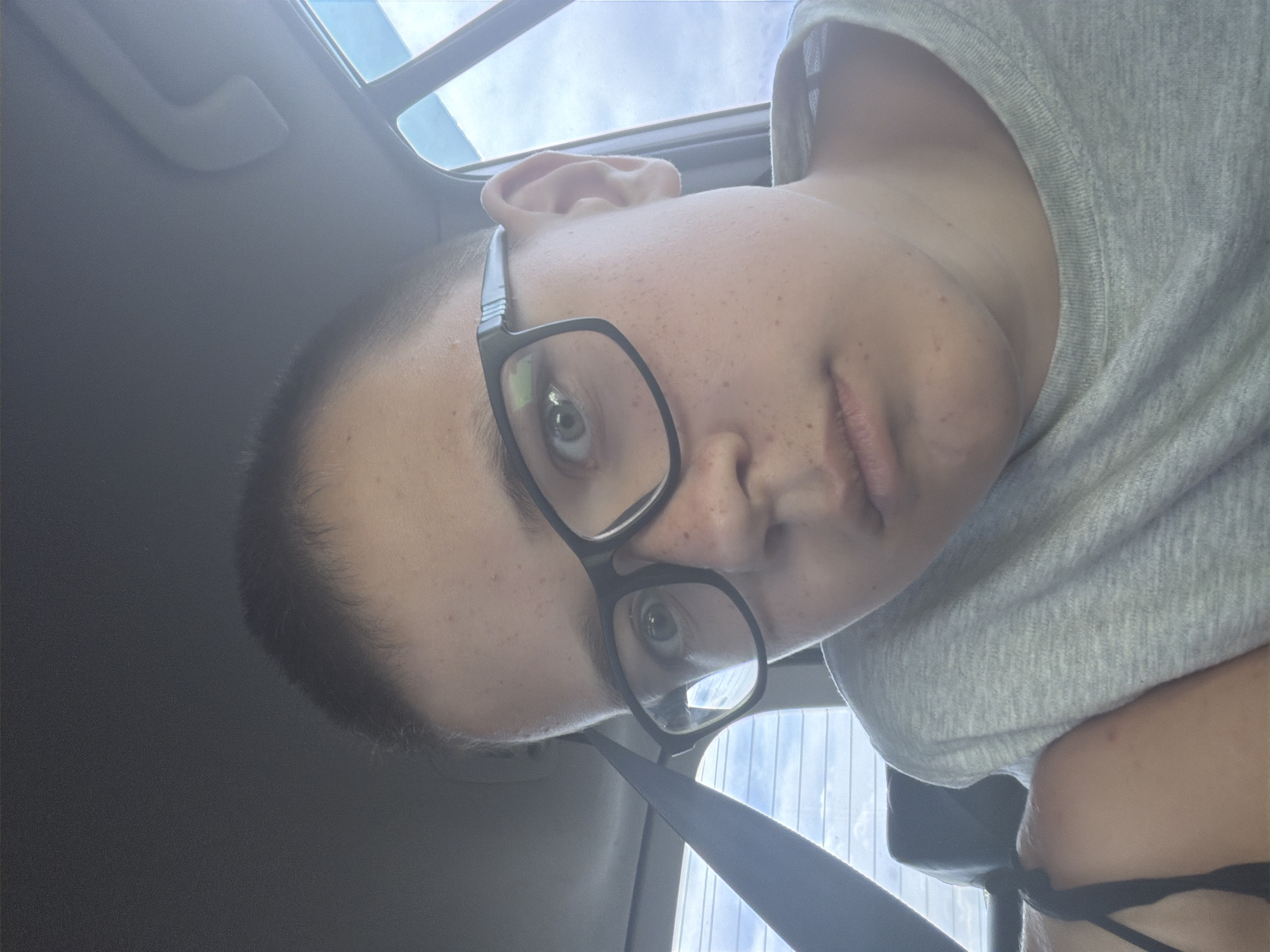 A boy, appearing to be in his early teens, is captured in a selfie taken from a car. He is wearing black-rimmed glasses and a light grey t-shirt. The background shows a car interior with a grey headliner and a portion of a window with white blinds, suggesting he is in transit. The lighting is bright, indicating it is daytime, and the sky outside the window appears overcast. The image does not contain any information that would place it in Teneevo, Russia.