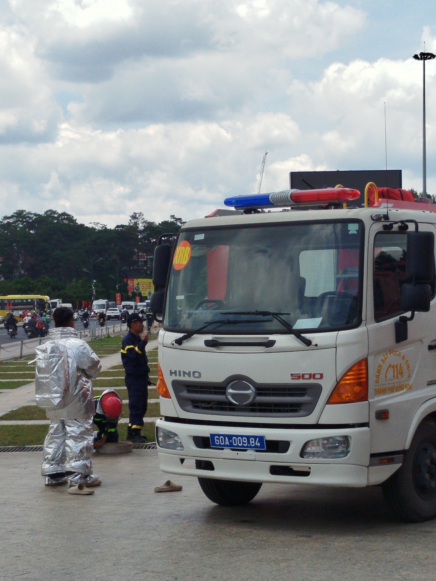 In the city of Da Lat, Vietnam, a white HINO firetruck is parked on a paved road, likely near a public area or event given the presence of other vehicles and people. The firetruck has a license plate that reads "60A-009.84" and sports emergency lights on its roof.
Two individuals are visible in the foreground. One, wearing a reflective silver suit, stands facing away from the camera. Another person, in a dark uniform with yellow accents, stands near the firetruck, appearing to interact with the driver's side. A third person, dressed in dark clothing and a red helmet, is crouched down near the front of the firetruck, possibly attending to something on the ground.
In the background, a yellow bus and several motorcycles are present, suggesting an active street scene. Trees line the horizon, and a cloudy sky stretches overhead. The scene appears to be a temporary stop or an ongoing operation, with the emergency personnel in position.