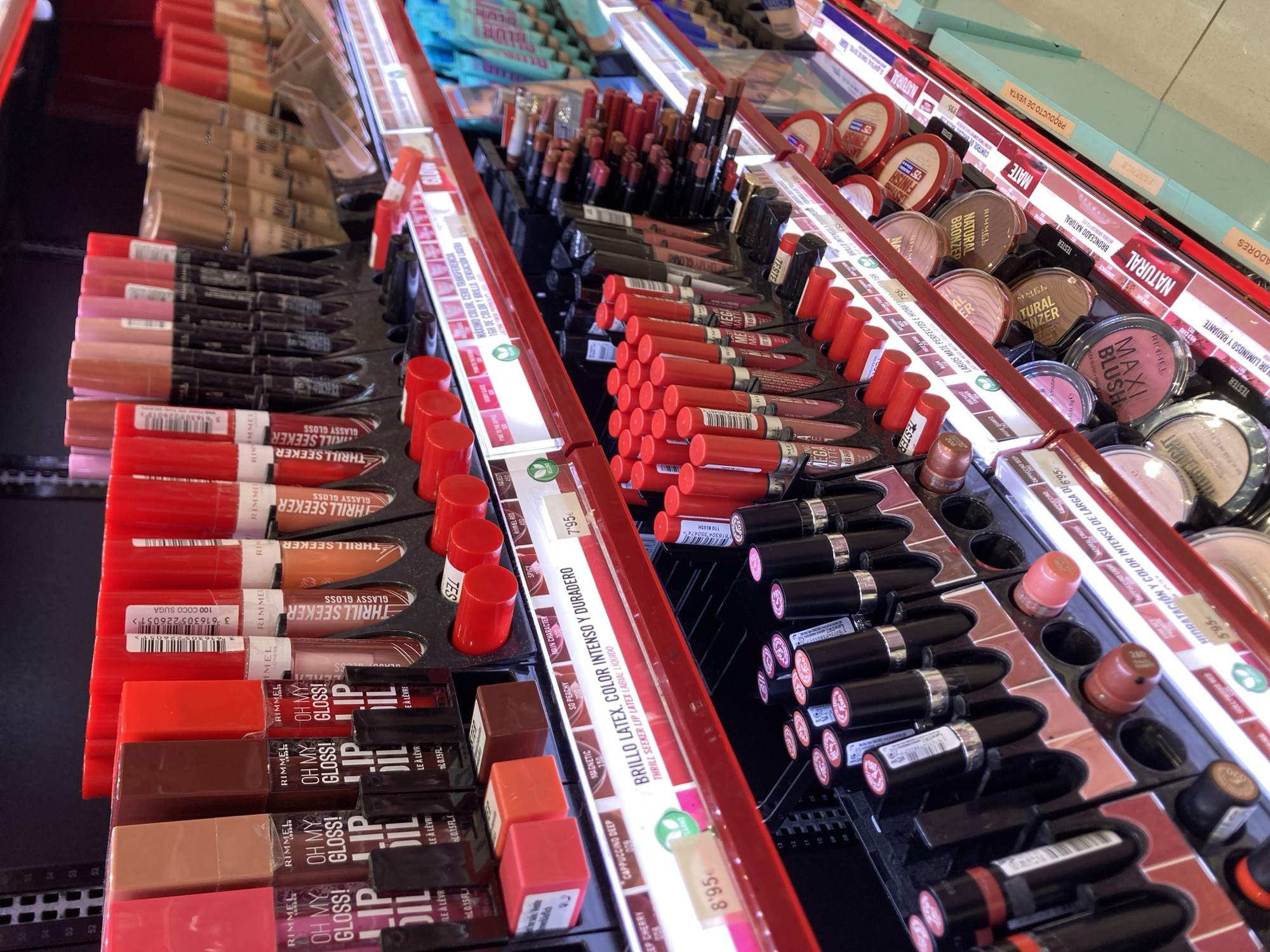 This is a retail display of makeup products, likely in a store in Barcelona, Spain. The image shows several shelves filled with various cosmetic items, primarily lipsticks and lip glosses. The products are arranged in an organized manner, with different brands and types clearly visible.
On the top shelves, there are numerous tubes of Rimmel London lip products, including "Oh My Gloss!" and "Thrill Seeker" lip glosses, in shades ranging from pinks and reds to nudes and browns. These are displayed in individual slots. Further down, other brands of lipsticks and lip liners are visible, also in a variety of colors.
The middle shelves feature more Rimmel London products, including "Brillo Latex," "Color Intenso y Duradero" lip glosses, and other lip products. There are also displays of pressed powders and bronzers, some with "Tester" labels. Price tags are visible on some of the items.
The lower shelves showcase more lipsticks and other makeup items. The overall environment appears to be a well-lit retail store with clean displays. The products are mostly new and in their packaging, suggesting a commercial setting. The presence of specific brands and product names, along with price tags and tester units, points towards a functioning retail environment where these items are available for purchase.