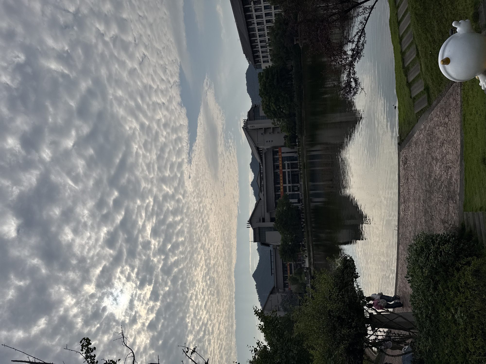 The photograph, taken in Jiangjin, China, shows a tranquil lake reflecting surrounding buildings and mountains. The sky is filled with a dense layer of altocumulus clouds. On the right side of the image, a grassy area and a paved walkway are visible, with a white decorative object resembling a duck placed at the edge. On the opposite side of the lake, buildings of varying heights, interspersed with trees, line the waterfront. In the foreground, foliage partially obscures a view of people near the water's edge.