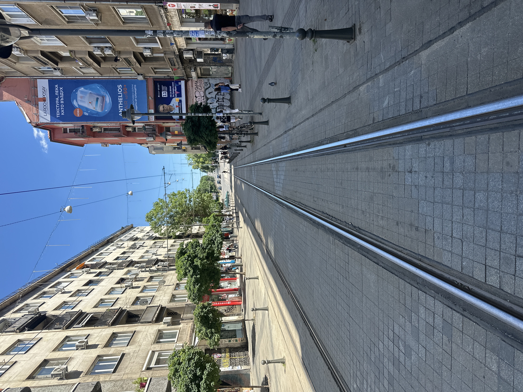 An upward-angle shot shows tram tracks running down the center of a paved street in a city. The tracks are flanked by sidewalks with lamp posts and trees. Buildings line both sides of the street, featuring balconies and air conditioning units. A billboard is visible on the right side of the street, advertising La Roche-Posay products. People are walking on the sidewalks and a pigeon is on the ground near a lamp post. Cars are seen in the distance at the end of the street. The sky is clear and blue.