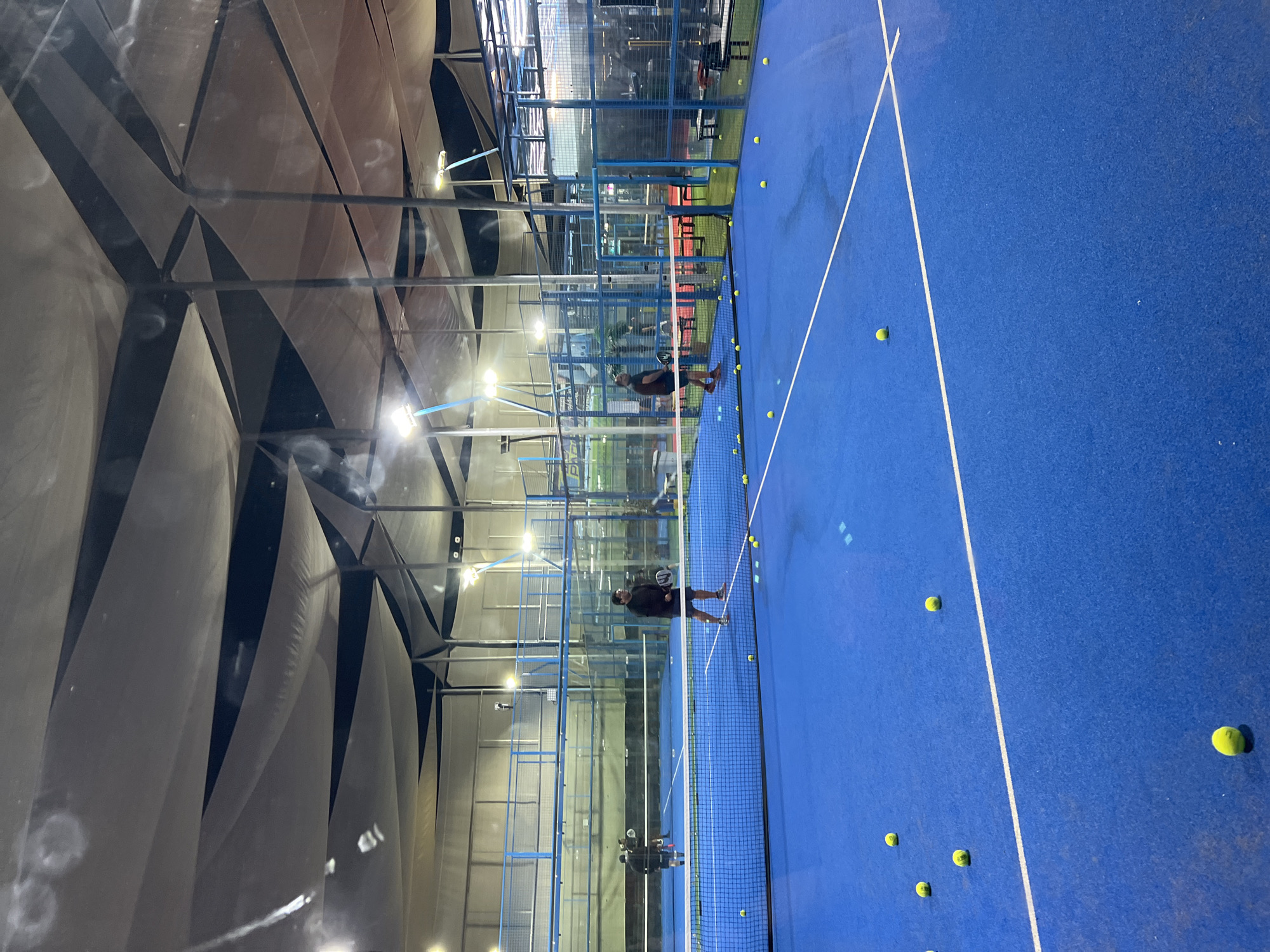 The image depicts an indoor padel tennis court in Arica, Chile. The court features a vibrant blue surface with white boundary lines. Two men are visible playing padel tennis. One man, wearing a dark shirt and shorts, is positioned on the right side of the court, holding a padel racket and facing the net. The other man, dressed in dark attire, is on the left side of the court, also with a racket, engaged in play. Several yellow padel balls are scattered across the court surface. The court is covered by a large, tensioned fabric roof structure, which is illuminated by artificial lighting. The perimeter of the court is enclosed by glass panels and metal fencing. The overall environment suggests an indoor sports facility.