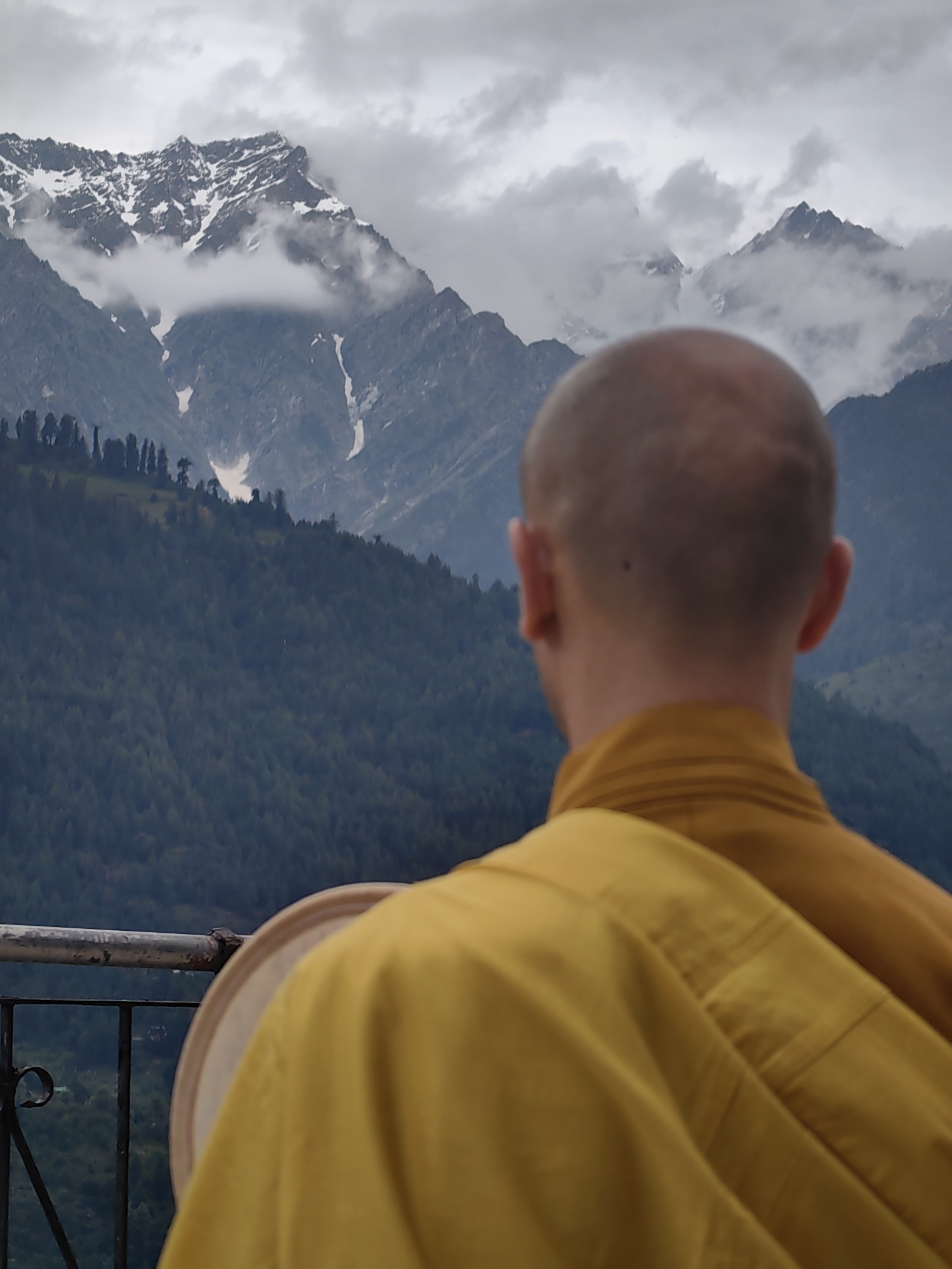The image captures a serene view from Vashisht, India, with a Buddhist monk in the foreground, his back to the camera, gazing at a majestic mountain landscape under a cloudy sky. The monk is wearing a saffron-colored robe. He is positioned on a balcony, with a railing and some dark metalwork visible. In his hands, he holds a circular object, possibly a drum, with a light-colored rim and a darker inner surface.
Behind the monk, a vast expanse of mountains stretches into the distance. The lower slopes are densely covered with dark green pine trees. Higher up, the mountains become rocky and rugged, with patches of snow visible on their peaks. The sky is overcast with dramatic clouds, contributing to the moody and contemplative atmosphere of the scene. The overall impression is one of tranquility and spiritual reflection amidst the grandeur of nature.