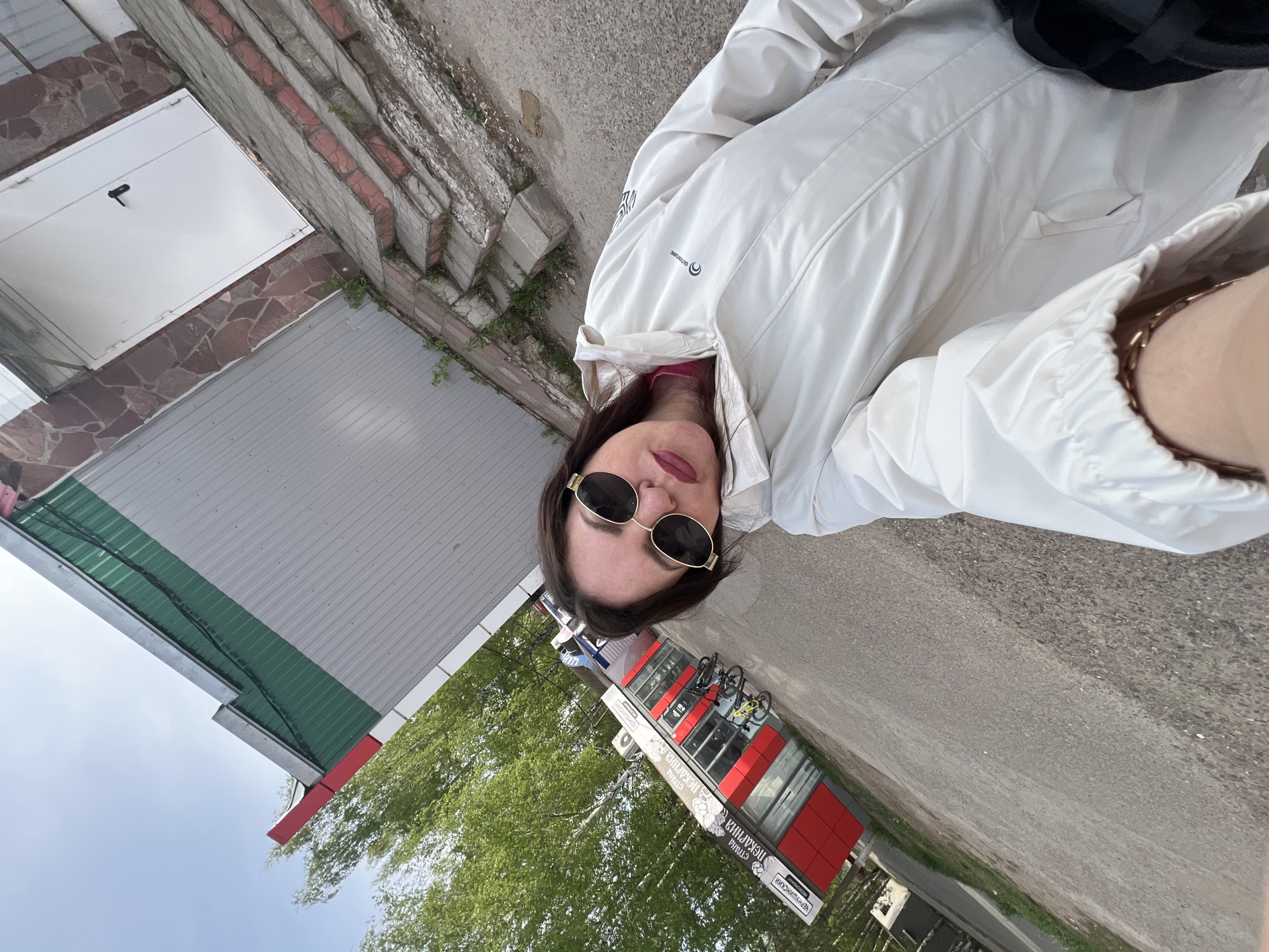 A woman with sunglasses and a white jacket is standing on a sidewalk in Noviy, Russia, near a building with a green and gray exterior. A red and white kiosk with the word "ПЕКАРНЯ" (bakery) in Russian is visible in the background. Bicycles are parked near the kiosk. A staircase with brick accents and some greenery is on the left of the image.