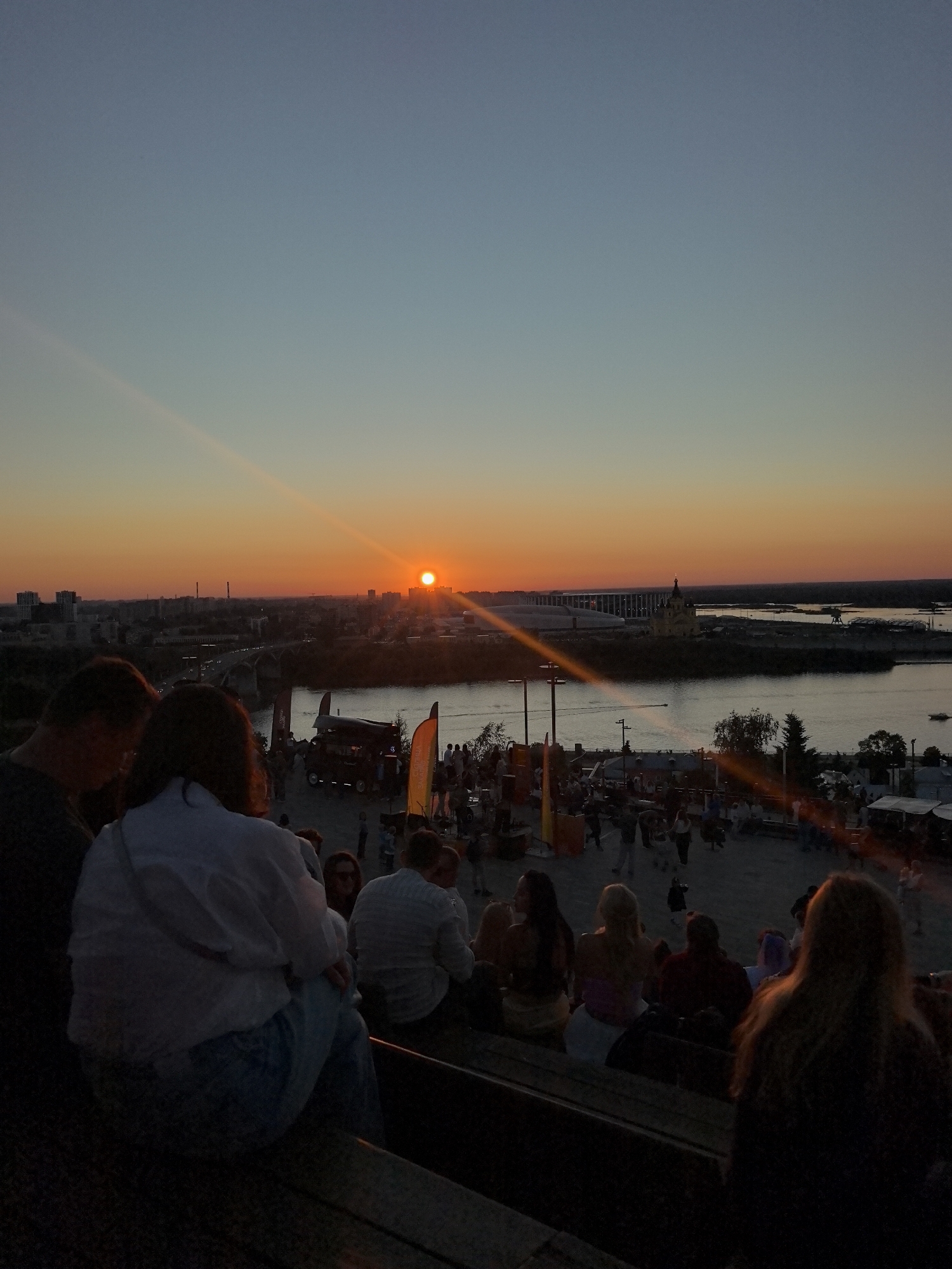 The image depicts a sunset over the city of Nizhny Novgorod, Russia, viewed from the Kremlin. The sun is low on the horizon, casting a warm orange glow across the sky and reflecting on the Oka River. In the foreground, a group of people are seated on bleachers, facing away from the camera, watching the sunset. They are casually dressed and appear to be enjoying the view.
In the middle ground, there is a gathering of people on a plaza, with food stalls and tents set up. This suggests an outdoor event or festival is taking place. Beyond the plaza, the Oka River stretches across the scene, with boats visible on its surface. The cityscape of Nizhny Novgorod is visible in the background, with various buildings and structures dotting the skyline.
The overall atmosphere is serene and peaceful, with the natural beauty of the sunset complemented by the vibrant activity of the city.