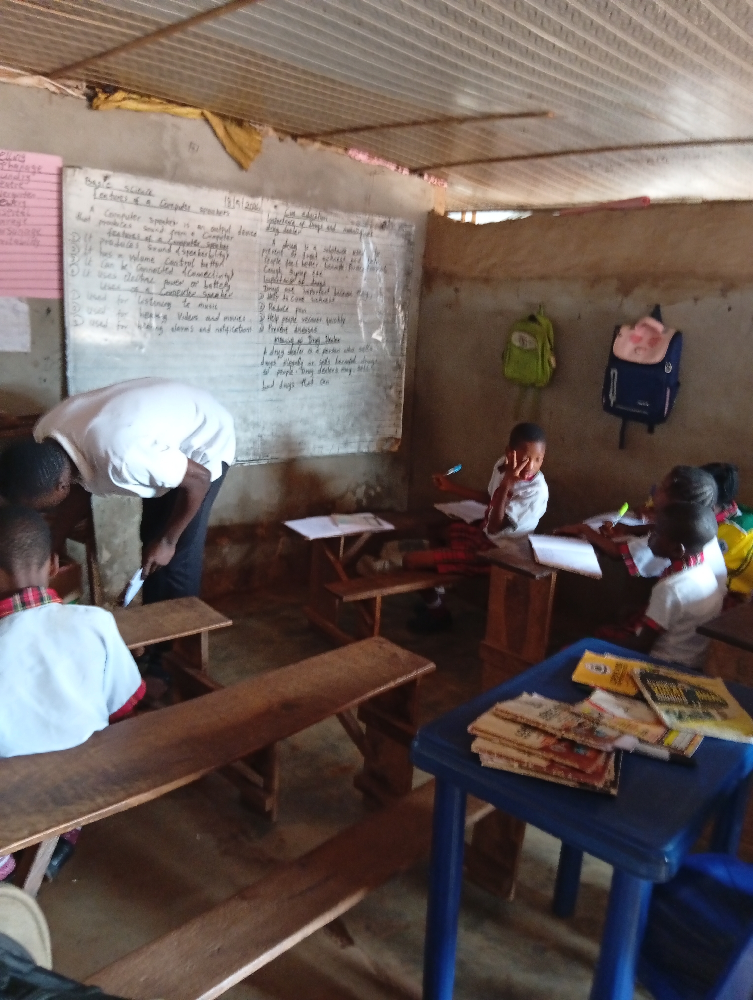 Here is a concise description of the image:
A classroom setting is depicted. A male teacher, wearing a white shirt and dark pants, is bending over near a student seated at a wooden bench. Several other students are at wooden desks, writing in notebooks. A whiteboard with handwritten text is on the wall behind the teacher. Two backpacks are hanging on the wall to the right of the whiteboard. There is a small blue plastic table in the foreground with books or papers on top. The room appears sparsely furnished and modestly constructed.