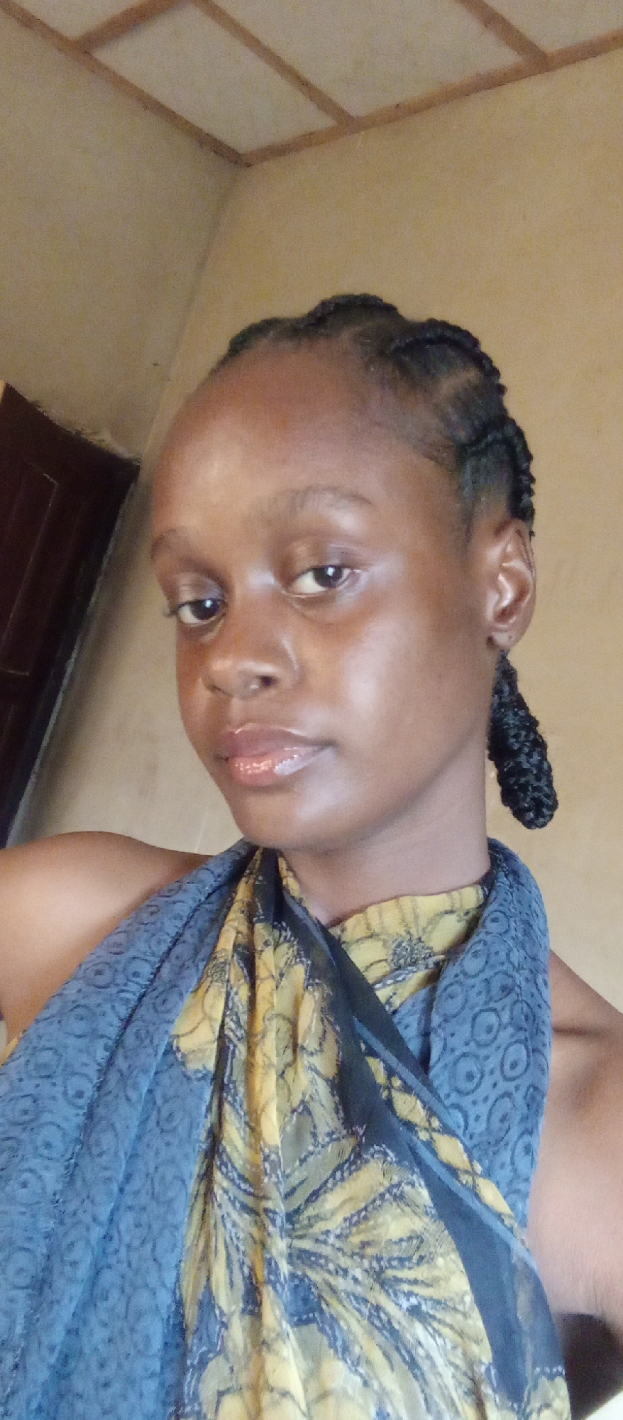 A close-up shot of a young woman with dark skin and braided hair, wearing a scarf with a blue and yellow pattern. The background appears to be an interior space with a pale yellow wall and a dark brown door in the distance. The location is not explicitly revealed as Akwanga, Nigeria from the image alone.