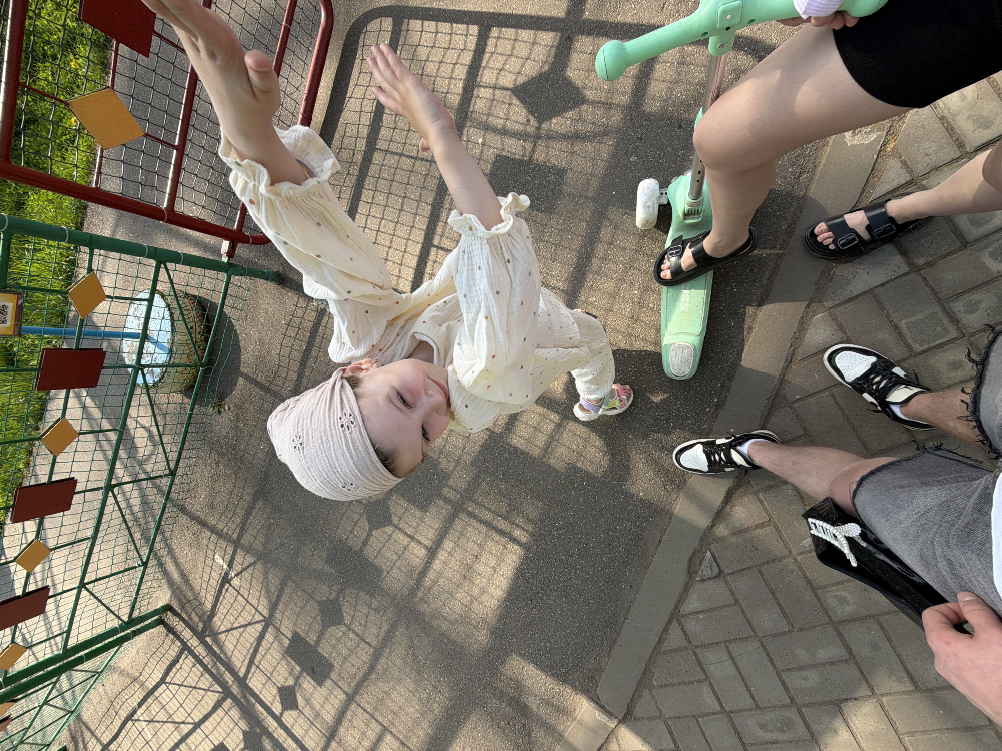 A low-angle, downward-facing photograph shows a young girl in a light-colored, polka-dotted, long-sleeved outfit and matching head covering, standing near a fence and gesturing with both arms raised. The fence is constructed of green and red metal bars. To the right of the girl, a green child's scooter is partially visible. Two individuals are also in the frame, with only their legs and feet visible. One person is wearing black shorts and black sandals, while the other is wearing grey shorts and white sneakers with black accents. The ground is a mix of asphalt and brick pavement, with shadows of the fence cast across the surface. The scene appears to be outdoors, possibly in a park or play area. This photograph was taken in Chaikovsky, Russia.
