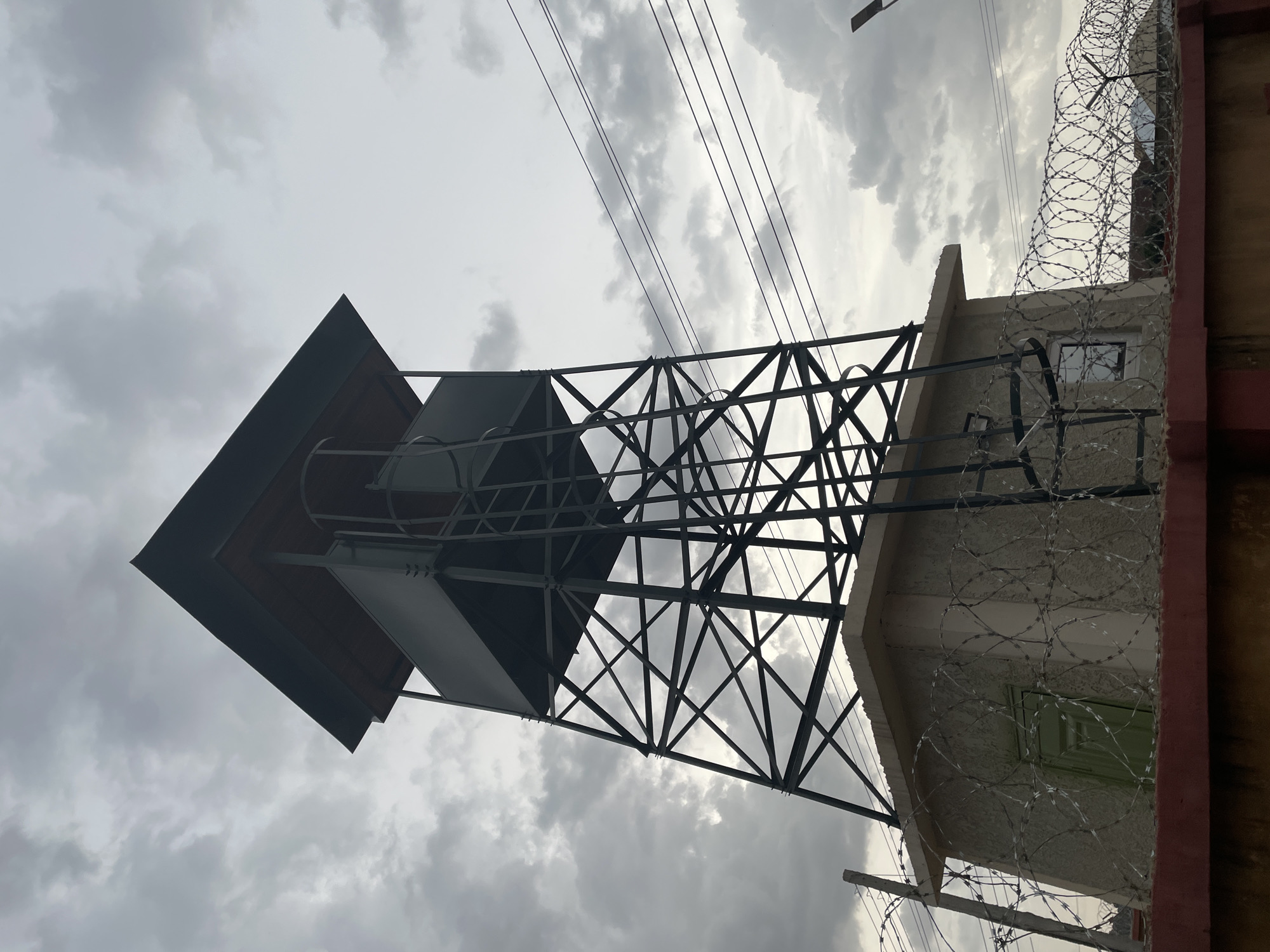 The image features a tall, metal observation tower with a square platform on top. The tower has a latticed framework. It is attached to a building constructed of concrete or stucco. The building is surrounded by barbed wire. The sky is overcast. Power lines run diagonally across the image. The location is Jos, Nigeria. No people are visible in the image.