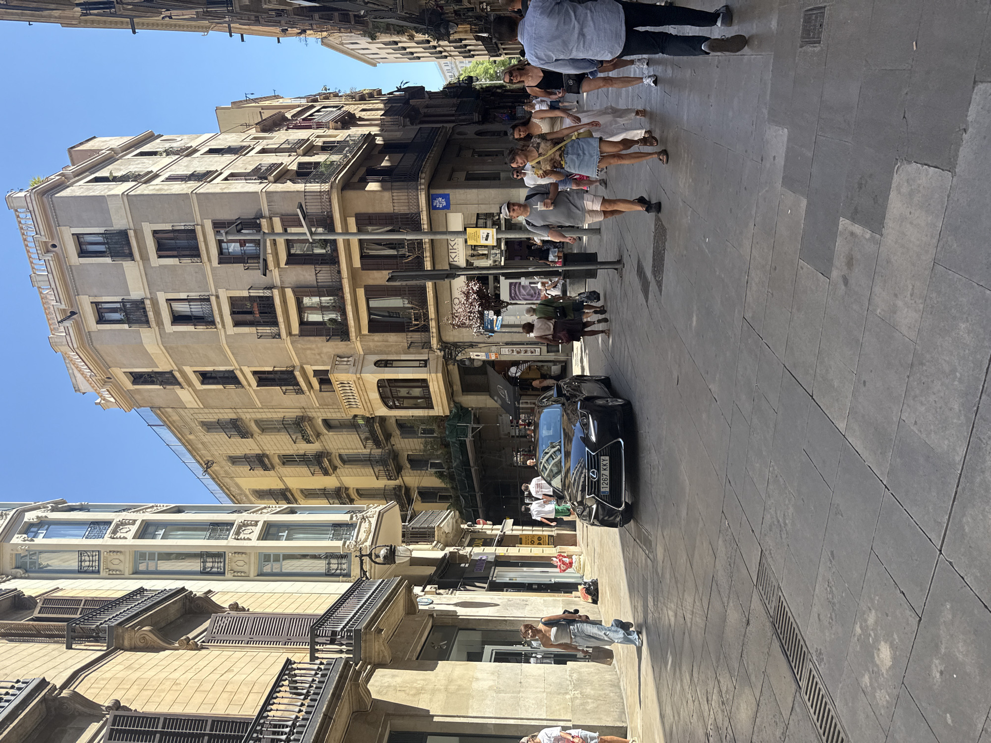 The image depicts a street scene in Barcelona, Spain, likely in the Gothic Quarter or El Born neighborhood, characterized by narrow, winding streets lined with historic buildings. A black Lexus sedan with license plate "1267 KKY" is the primary subject in the foreground, moving along the cobblestone street. Several pedestrians are visible, walking on the sidewalk or crossing the street. On the left, a woman in a grey tank top and jeans walks past a store with large windows. On the right, a group of people are walking towards the viewer. The buildings flanking the street are multi-story structures with traditional architecture, featuring balconies with wrought-iron railings and shuttered windows. Sunlight casts shadows on the ground, indicating a bright, sunny day. The overall atmosphere is that of a bustling European city street during daytime.
