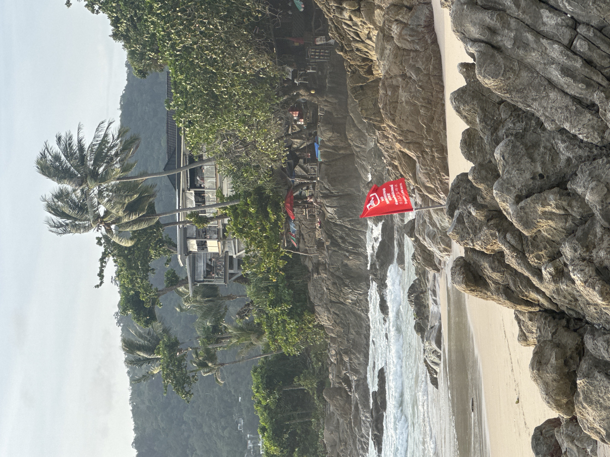 This image depicts a rocky beach in Patong, Thailand, with rough waves crashing against the shore. In the foreground, large, textured rocks dominate the scene, with a small stretch of sandy beach visible behind them. A bright red flag is planted firmly in the sand, bearing text in English and possibly Thai, indicating "NO SWIMMING HERE."
Behind the rocks and beach, a more developed area is visible. Several palm trees with wind-swept fronds are scattered across the landscape. A multi-story building, possibly a restaurant or resort, sits on a higher elevation, with balconies overlooking the ocean. People are visible on the balconies, suggesting activity within the building. Further back, lush green hills form the backdrop, with a hazy sky overhead. Small structures and umbrellas are also present on the rocky coastline, hinting at beachside services or amenities. The overall atmosphere suggests a slightly overcast day with strong winds, as evidenced by the moving palm fronds and the choppy sea.