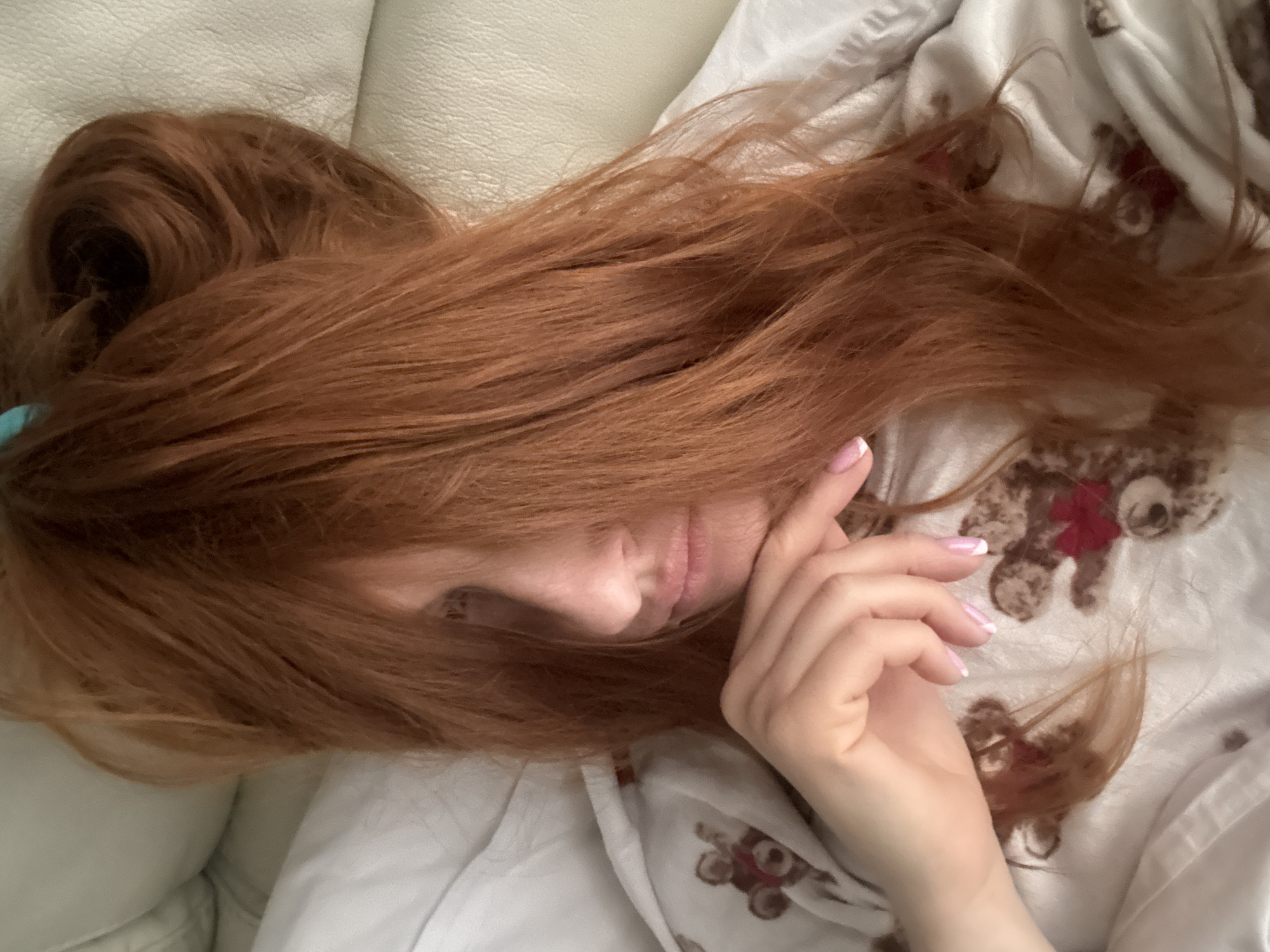A woman with long, red hair rests on a white leather surface, partially covered by a white blanket patterned with teddy bears. Her face is partially obscured by her hair, with her right hand resting near her cheek. The location is unspecified and unidentifiable in the image. There is no indication of the city of Budva, Montenegro, being present.