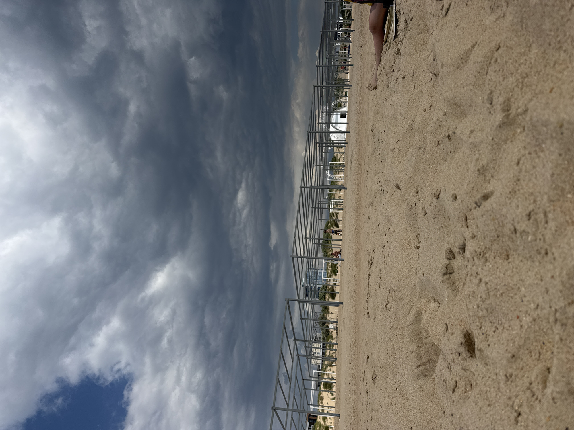 The image depicts a cloudy day at a beach in Vityazevo, Russia. The foreground is dominated by sand with footprints. In the mid-ground, a series of metal structures, presumably for beach umbrellas or cabanas, are erected in rows. A few people are visible in the distance, some standing near the structures. Beyond the structures, there are some buildings and sand dunes. The sky is filled with dark, heavy clouds, suggesting an impending storm. In the right foreground, a person's legs are visible, lying on the sand. The overall scene is one of a beach preparing for or experiencing inclement weather.
