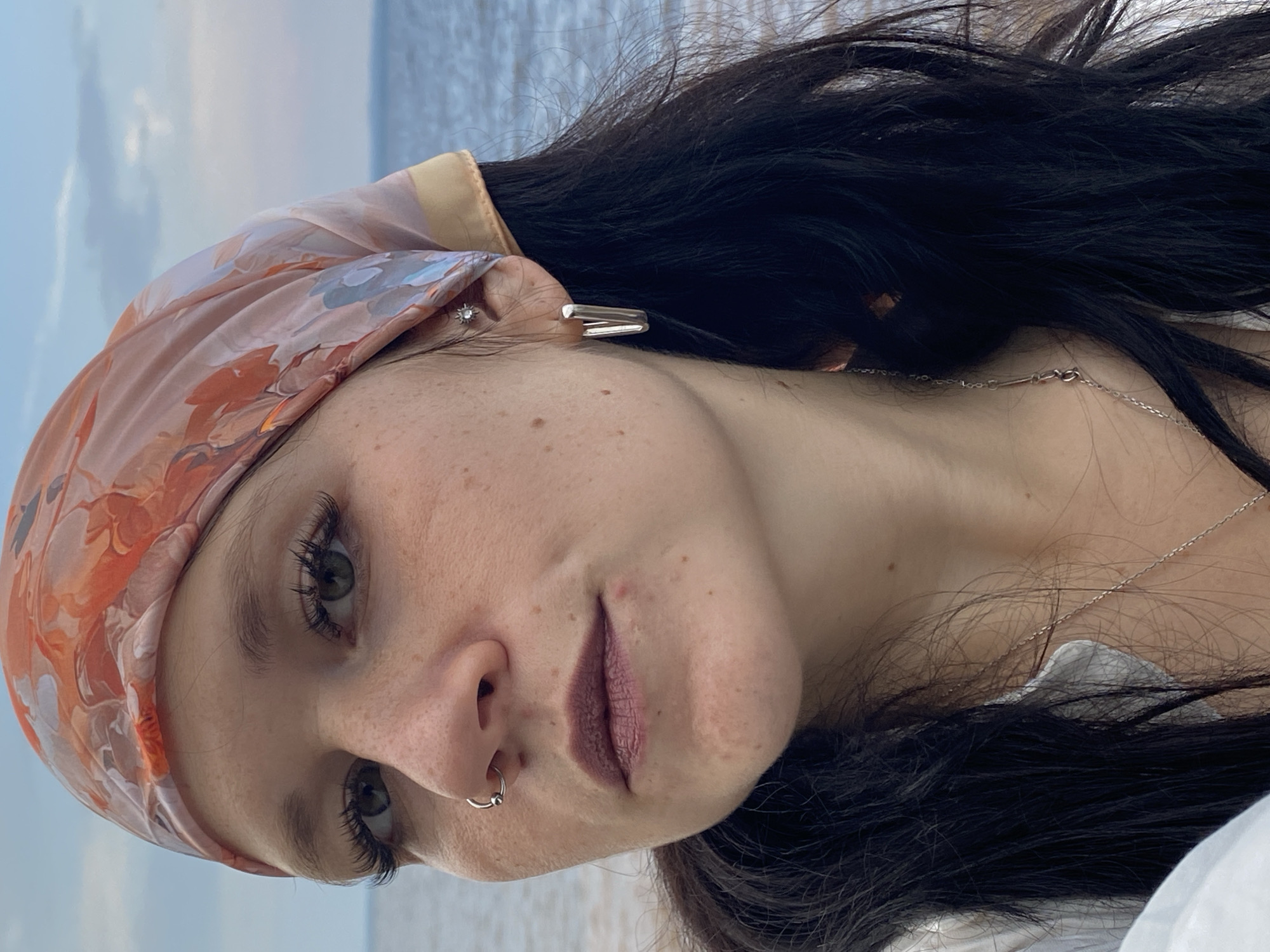 A close-up, waist-up outdoor portrait shows a woman with dark hair wearing a light-colored patterned headscarf and a white shirt. She has a nose ring and earrings. The background is a body of water under a cloudy sky, likely a bay or lake. The lighting suggests it is late afternoon or early evening. The woman is looking slightly away from the camera. The overall scene conveys a casual, relaxed atmosphere.