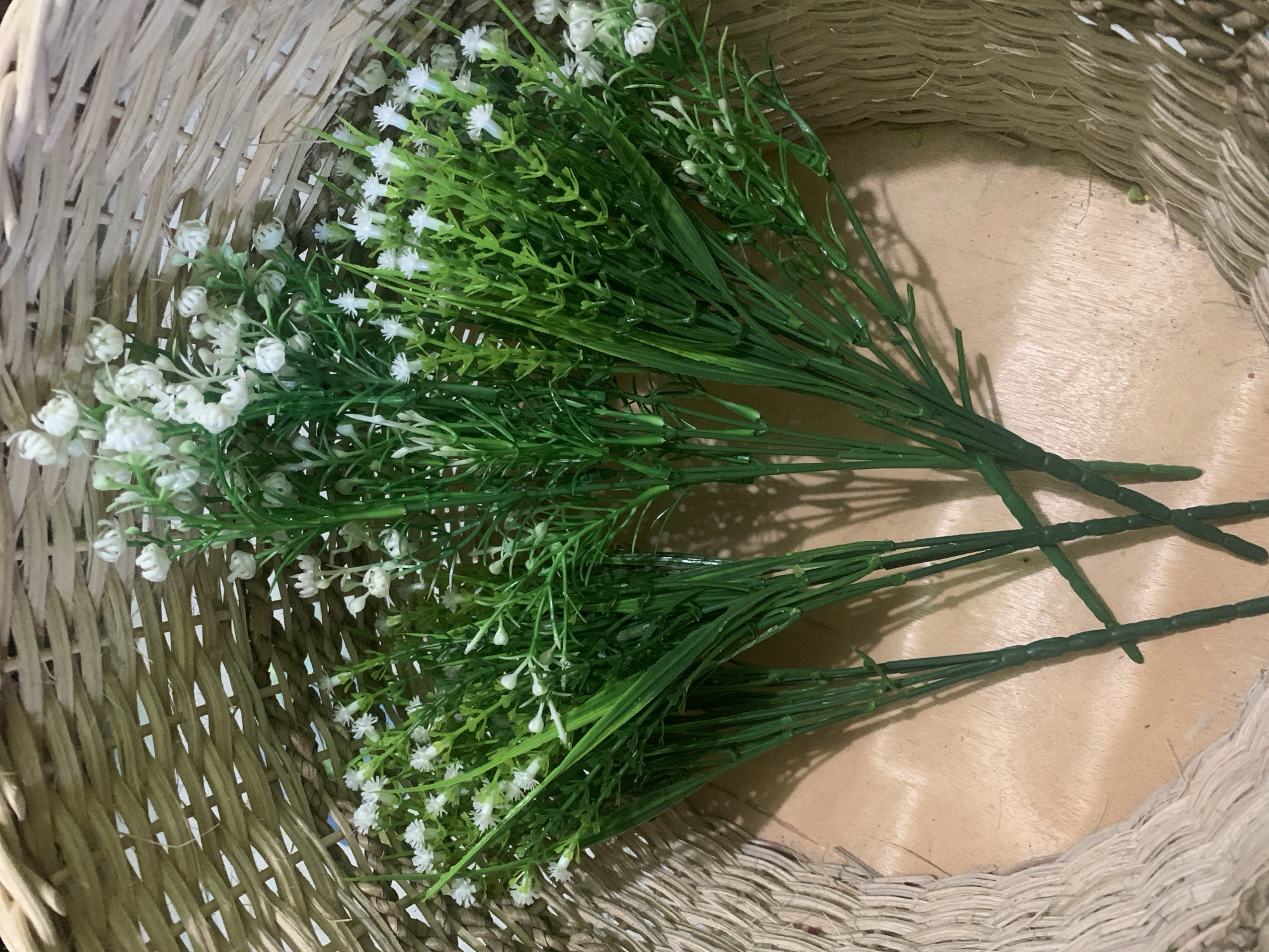 In Kuala Pembuang, Indonesia, a close-up, overhead shot captures a woven basket filled with artificial flowers and greenery. The basket, made of natural fibers, has a rough texture and is predominantly beige. Inside, several bundles of faux plants, featuring numerous small white blossoms and slender green leaves, are arranged together. The lighting casts soft shadows on the wooden surface beneath the basket. The scene is static, with no visible individuals or actions. The environment is domestic or decorative, emphasizing the handcrafted appearance of the basket and the artificial nature of the floral arrangement.