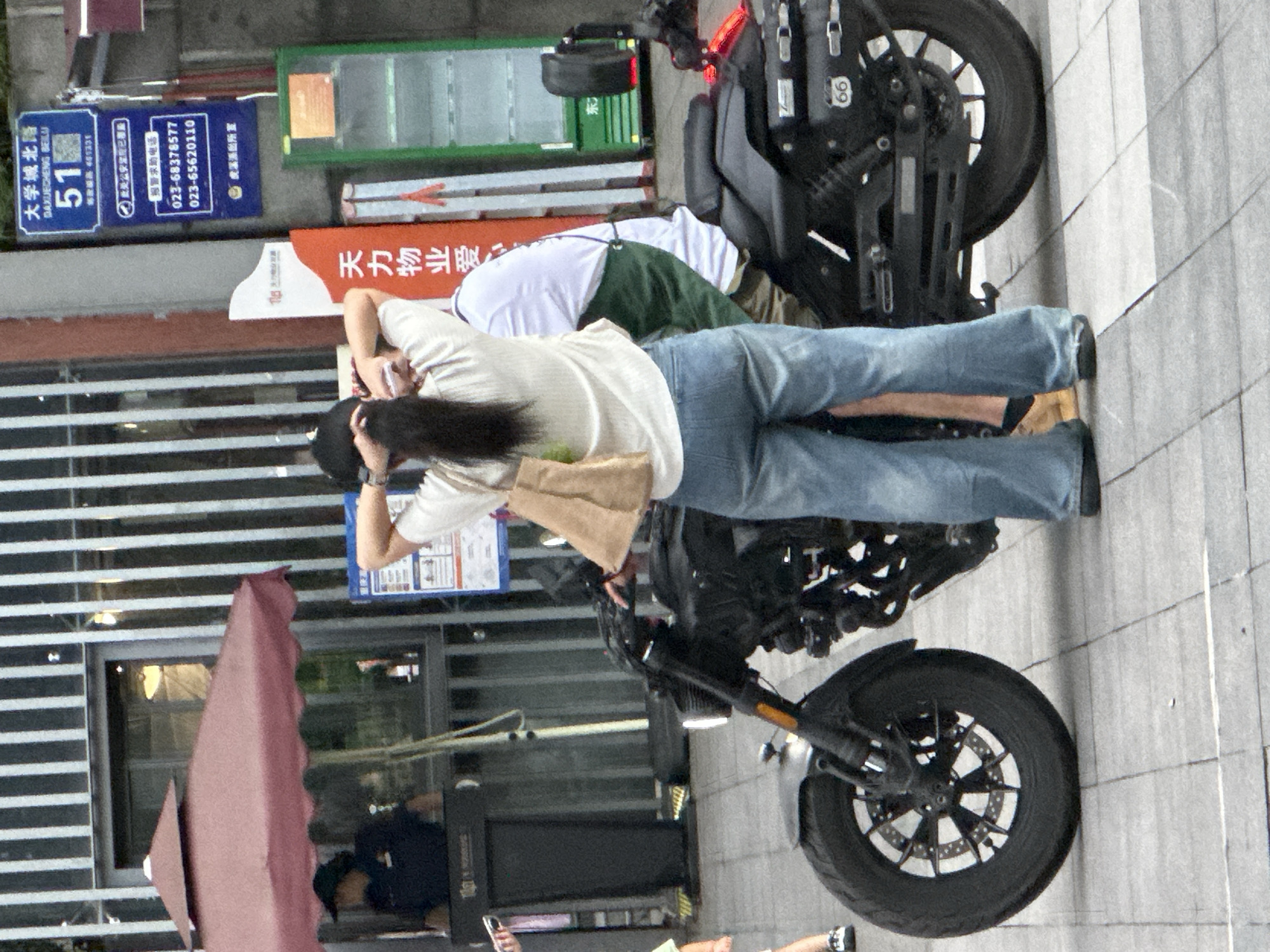 This is a vertical, medium-shot photograph taken outdoors. The scene appears to be a sidewalk or paved area next to a building, possibly in an urban setting given the presence of signage and a parked motorcycle.
The primary subjects are two individuals. One person is leaning forward, with their head down and arms extended, appearing to be looking at something on the ground or a device in their hands. They are wearing a light-colored long-sleeved shirt and blue jeans. Their hair is dark and long, falling over their face. The second person is partially visible, wearing a white shirt and a green apron or vest, and their body is angled away from the camera.
A black motorcycle with a modern design is parked in the center of the frame. It has a prominent front wheel and suspension system visible. A portion of a pink, tent-like structure is on the left side of the image, partially obscuring a building with horizontal blinds.
Several pieces of signage are visible. One blue sign features Chinese characters and numbers, including "大学城北路 DAXUECHENG BEILU" and "51". Another sign, red and white, has Chinese characters that appear to be a company name, "天力物业爱". A green cabinet-like structure is also visible in the background, possibly part of the building's facade. The ground is paved with light-colored tiles.
The time of day is unclear, but the lighting suggests it might be daytime. The overall atmosphere appears to be casual and everyday. The visible text provides location context, indicating "Daxuecheng North Road" and a police station ("虎溪派出所室").