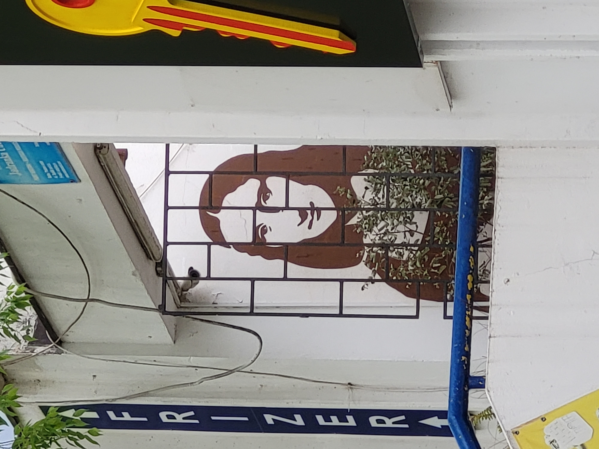 This sideways-oriented image captures a street scene in Budva, Montenegro, under bright daylight. In the upper portion, a large, stylized yellow key with red accents is prominently displayed against a dark background, likely signifying a locksmith or key-cutting business.
Below this, a significant part of the frame is dominated by a graphic or mural on a white wall. This artwork features a minimalist portrait of a person's face, possibly a woman, with dark brown hair and a neutral expression, gazing towards the right. A dark metal grid partially covers the lower section of the face and extends across the wall. Green foliage and branches are intertwined with the grid and the mural, introducing a natural element.
Architectural details include white concrete or plaster structures forming an overhead beam or ceiling element, across which thin electrical wires are draped. On the upper left, a small, partially obscured blue sign with white text begins with "Jastranka G...". A vertical blue pole is visible on the right side of the frame. At the very bottom, a dark blue band with white letters reads "R E Z I R F". When viewed correctly (rotated upright), this text spells "FRIZER," indicating the establishment is a hairdresser or barber shop. The scene captures the decorated exterior of these commercial premises, with no specific activity taking place.