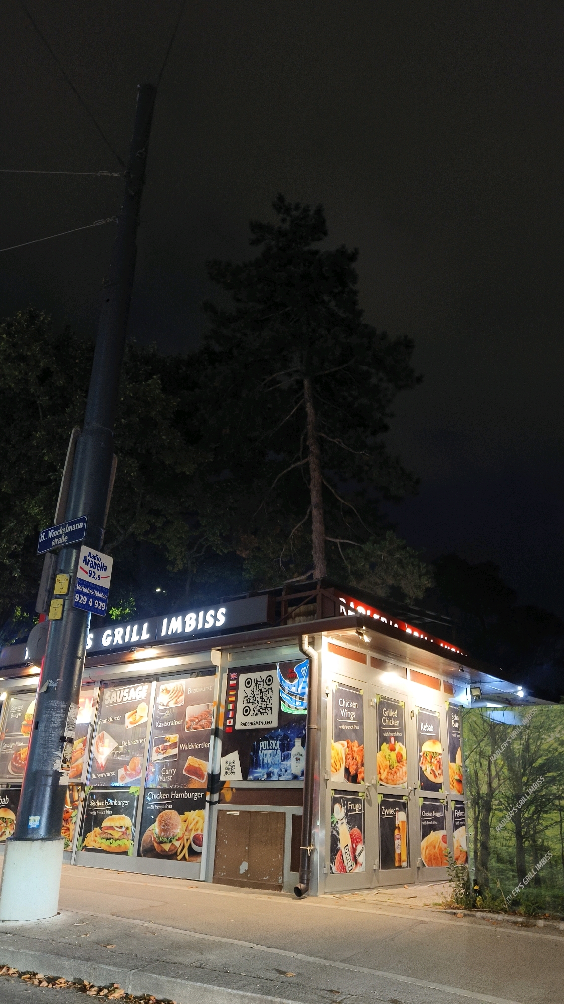 The image captures a street scene at night in Vienna, Austria. A long, dark lamppost stands prominently in the foreground, with street signs attached to it. One sign indicates "5. Winckelmann straße," and another displays "Radio Arabella 91,9" along with a phone number. Adjacent to the lamppost is a food stand, identified by a large illuminated sign that reads "GRILL IMBISS."
The food stand is adorned with numerous advertisements for various food items, including sausages, hamburgers, chicken wings, and kebabs, each accompanied by images of the food. Some of the signage also displays QR codes and text in German and English. The stand is illuminated by overhead lights, creating a well-lit area despite the nighttime setting. In the background, a tall pine tree rises above the food stand, its branches reaching into the dark sky. The overall environment suggests a casual dining establishment in an urban setting.