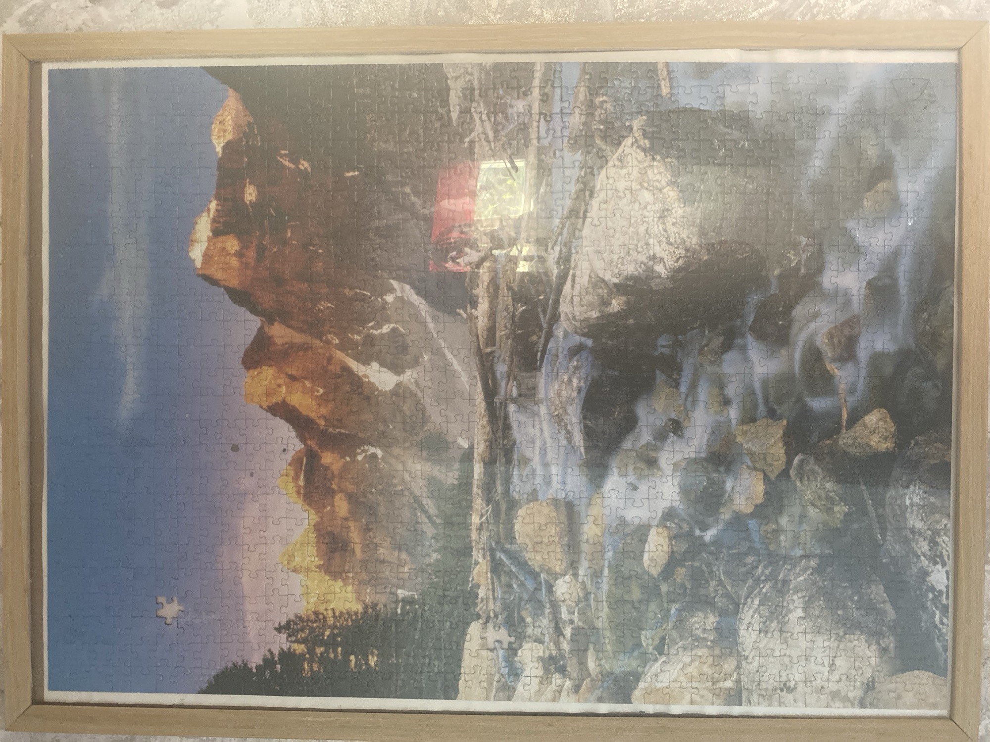 The image displays a framed jigsaw puzzle depicting a mountain landscape. The puzzle shows rugged, sunlit peaks in the background with a rocky riverbed in the foreground. A small piece of the puzzle is missing from the upper left quadrant. The scene is illuminated by natural light, likely daylight, creating warm tones on the mountains and a cooler palette in the river area. There are no people or animals visible in the puzzle's image. The puzzle is framed by a light-colored wooden frame. The puzzle is positioned on a textured surface. There is no indication or information suggesting the image is related to the city of Blagoevgrad, Bulgaria.