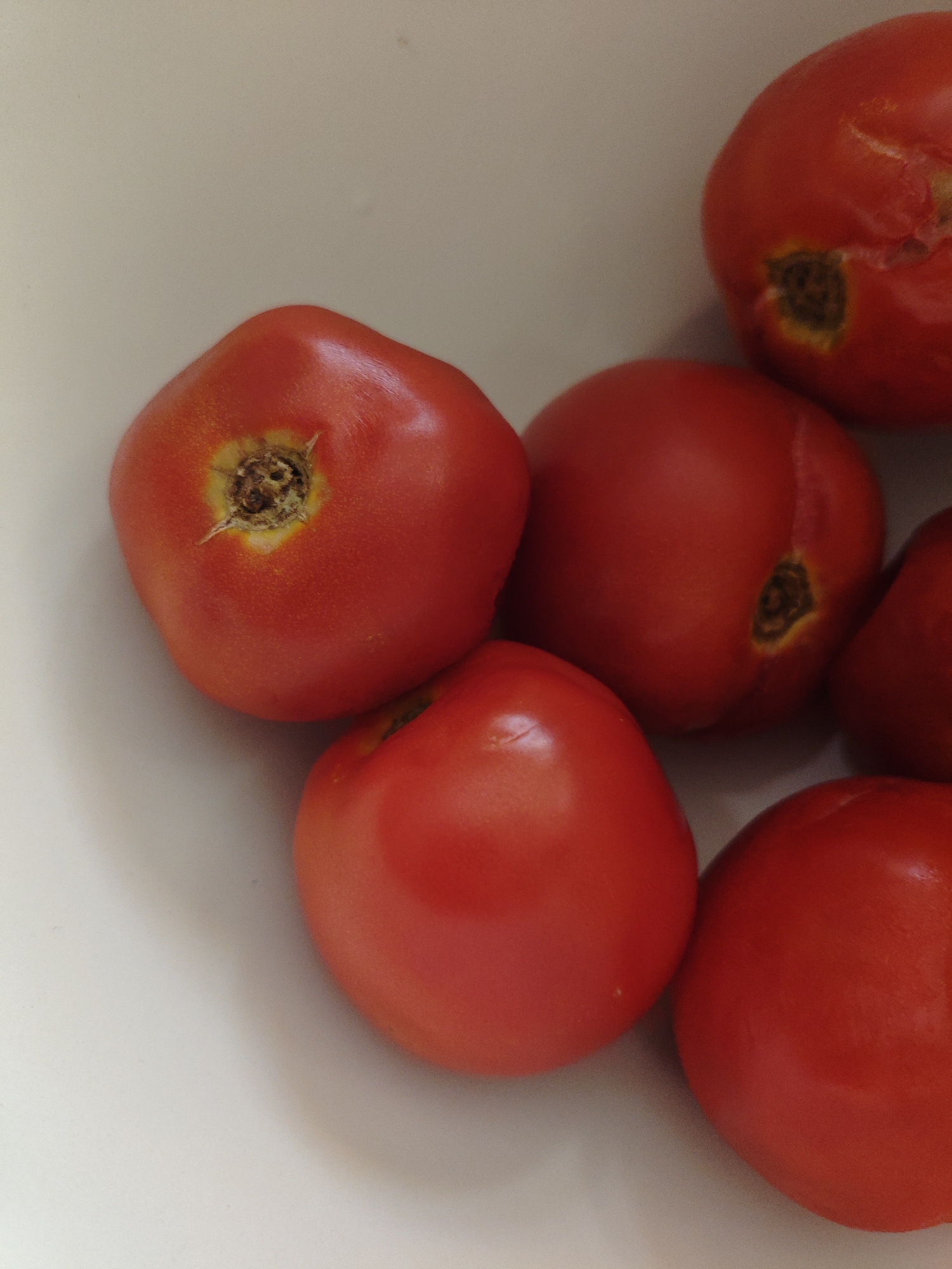 This is a close-up, top-down shot of several ripe red tomatoes in a white bowl. The tomatoes are the main subject, filling most of the frame. Some tomatoes have a visible stem end with a rough, brown texture. The lighting is bright and even, highlighting the smooth, curved surfaces of the tomatoes. The background is a clean, white surface, likely the inside of the bowl. There are no people or any indications of location within a city, including Armavir, Russia.
