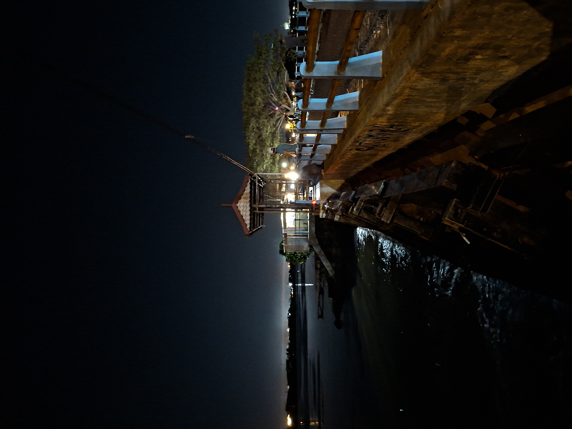 This is a night photograph taken from a pier looking out over the water. In the foreground, a concrete barrier lines the edge of the pier, with white pillars and wooden railings visible. The water below is dark and choppy, reflecting some of the lights from the shore and a gazebo structure further down the pier. Several people are standing on the pier, some near the gazebo, and others further along the railing. A large, leafy tree is visible in the background, illuminated by lights. The sky is dark, with distant city lights visible on the horizon. The location is identified as the Tugu Rencong monument area in Lhokseumawe, Indonesia, a popular spot for locals to gather and enjoy the waterfront.