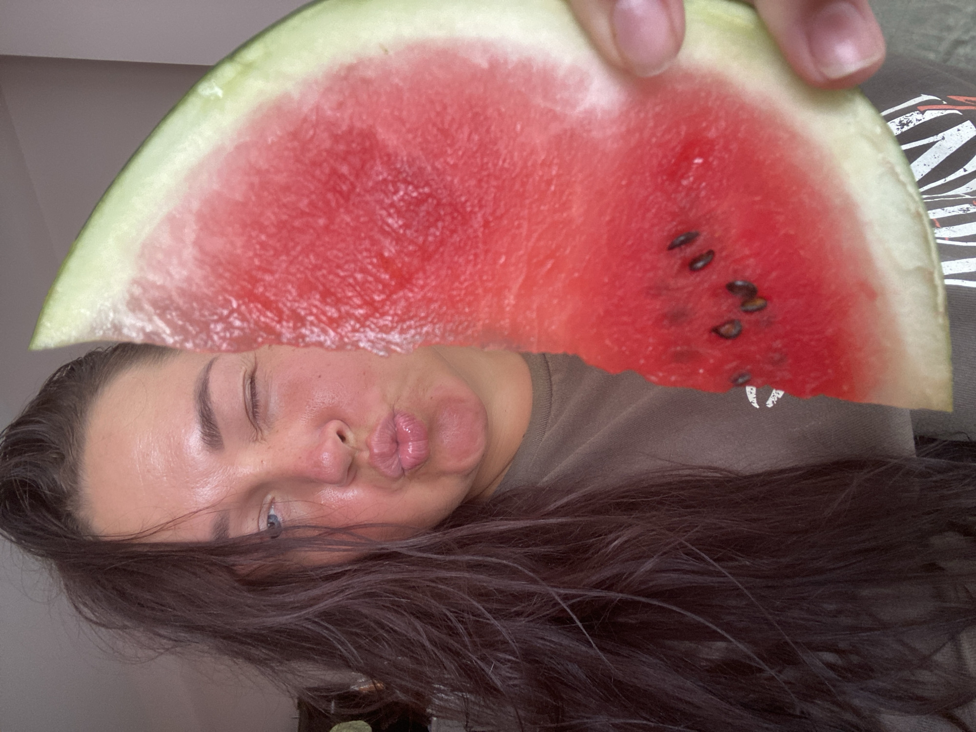 The image shows a close-up, selfie-style shot of a woman holding a slice of watermelon in front of her face. She has dark, long hair that is partially obscuring her face. Her left eye is closed in a wink, and her lips are puckered. The watermelon slice is large and vibrant red, with visible seeds. She is wearing a dark-colored t-shirt. The background is neutral and out of focus, suggesting an indoor setting. There is no indication of the city of Sochi, Russia, in the image.