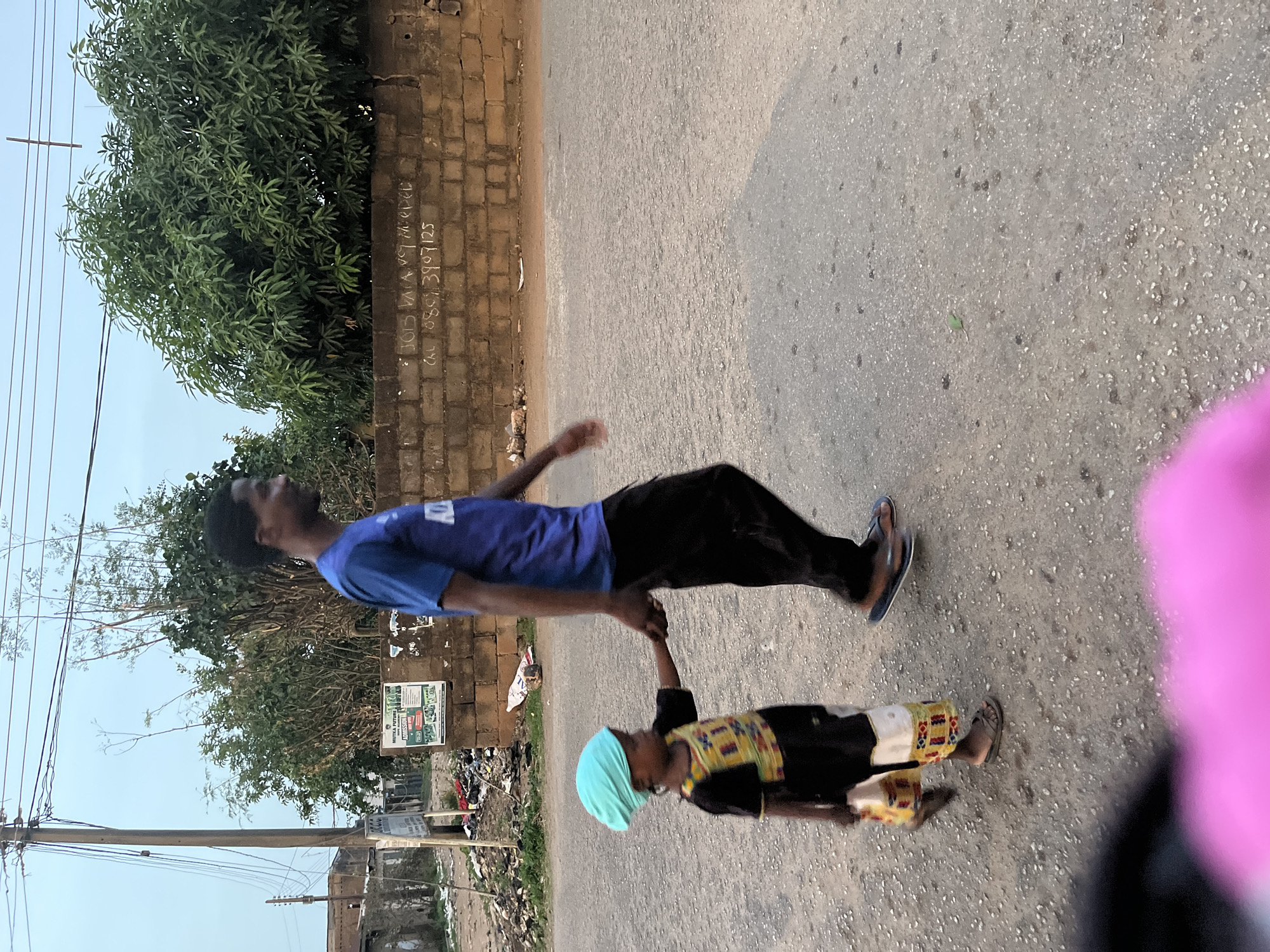 A street scene in Jos, Nigeria features an adult male wearing a blue shirt and black pants walking alongside a child. The child is wearing a light blue head covering, a dark shirt with a patterned yellow and red vest, and tan pants. They are holding hands as they walk on a grey paved surface, likely a road or sidewalk. Behind them, there's a wall covered in greenery and what appears to be a brick building. Litter is visible beside the wall. Power lines are overhead. The area is urban with some vegetation. A pink object obscures part of the foreground.