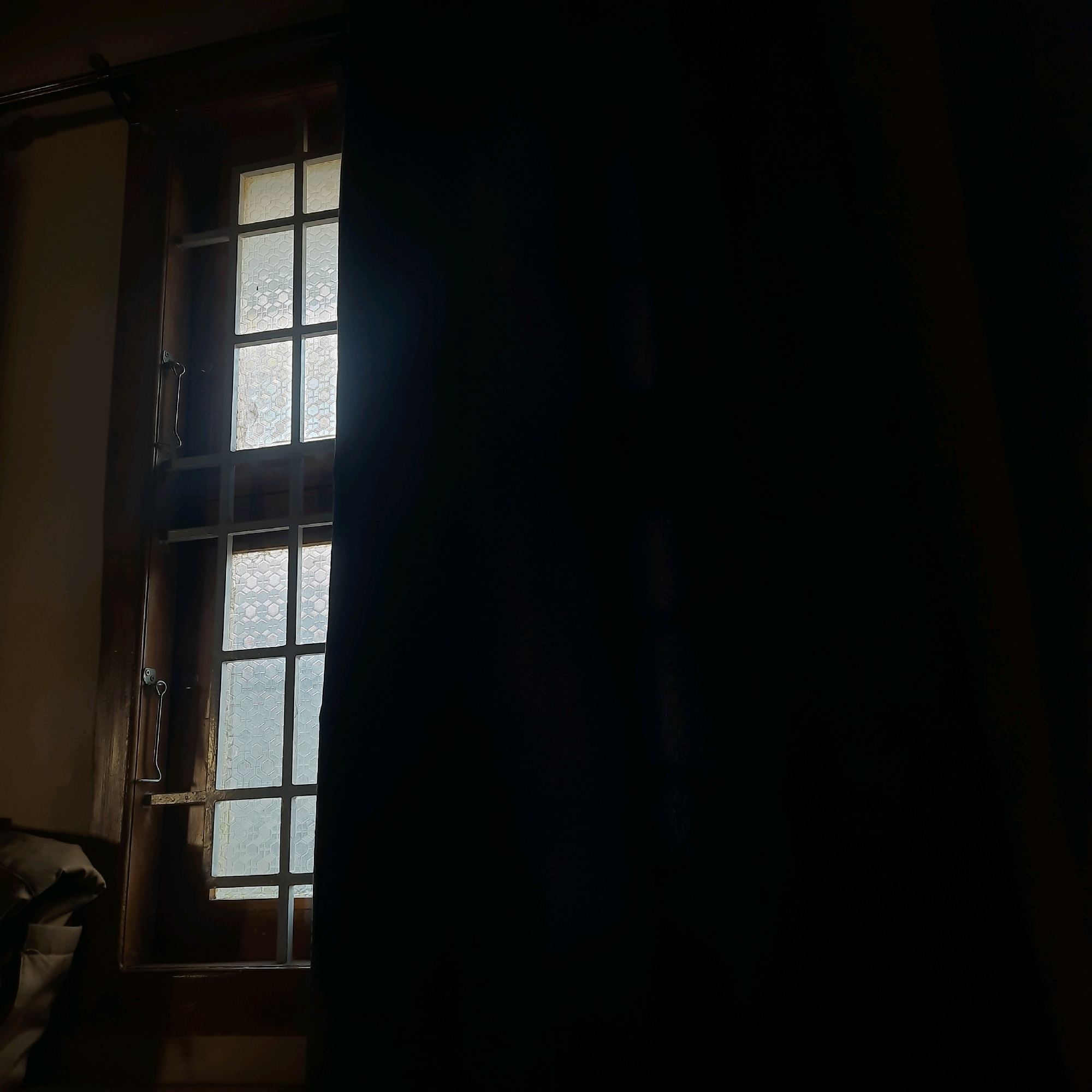 The image shows a window with a dark curtain partially obscuring the view. The window has a wooden frame and a metal grate. The glass in the window is frosted with a geometric pattern. Light is visible through the window, suggesting it is daytime outside. A metal latch is attached to the wooden frame. To the left of the window, a portion of what appears to be a folded piece of fabric, possibly clothing or bedding, is visible in the lower-left corner. The overall scene is dimly lit, with the curtain creating a strong contrast between light and shadow. The image is undated and does not provide any specific information about its location in Coimbatore, India, nor does it depict any people or actions.