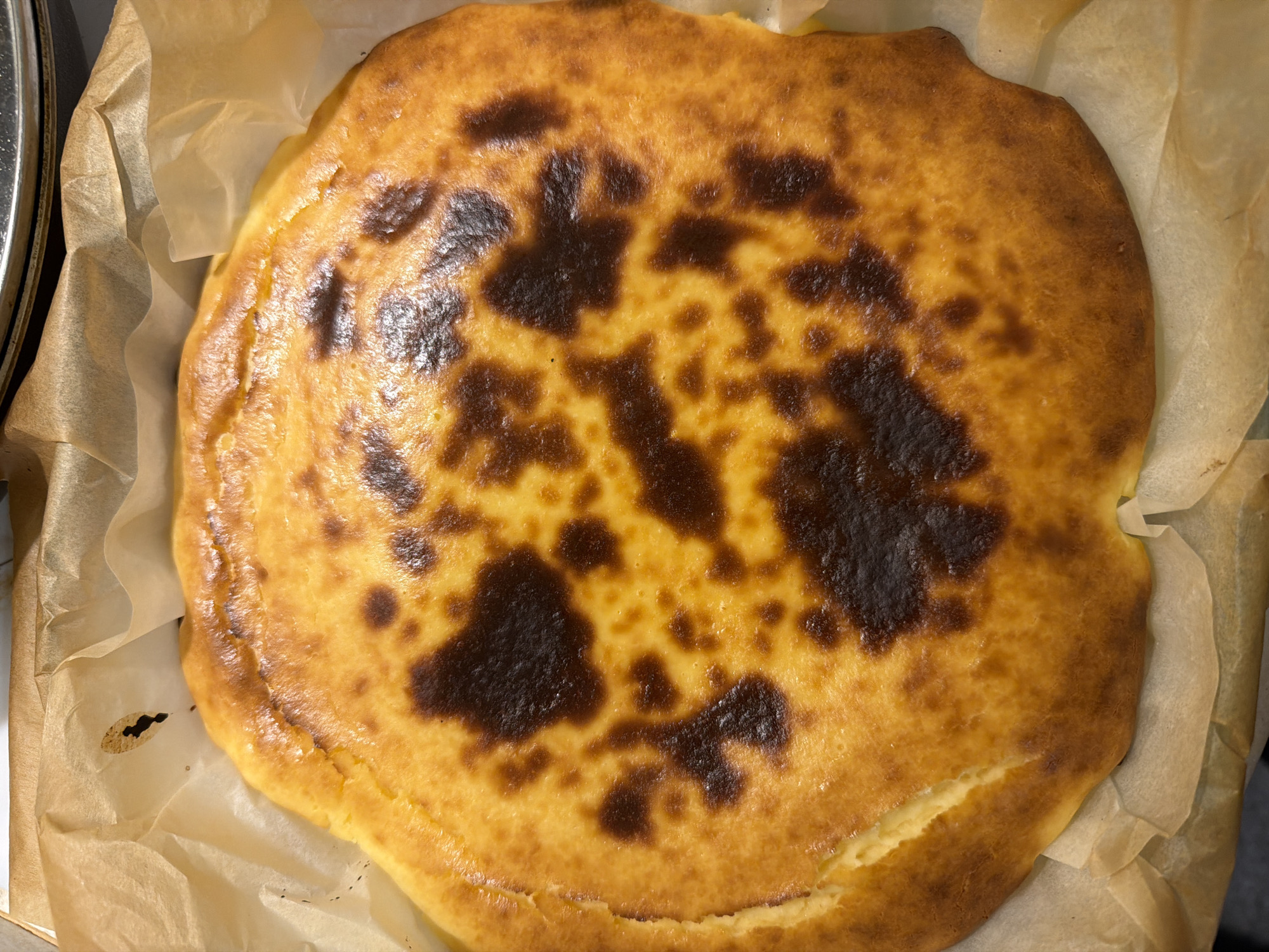 The image shows a baked Basque cheesecake on parchment paper. The cheesecake has a browned and cracked top with dark spots. The cheesecake is roughly circular in shape. The parchment paper appears wrinkled and covers a metallic surface. The image does not contain any information about the location in the city of Budva, Montenegro, people, actions, or interactions.