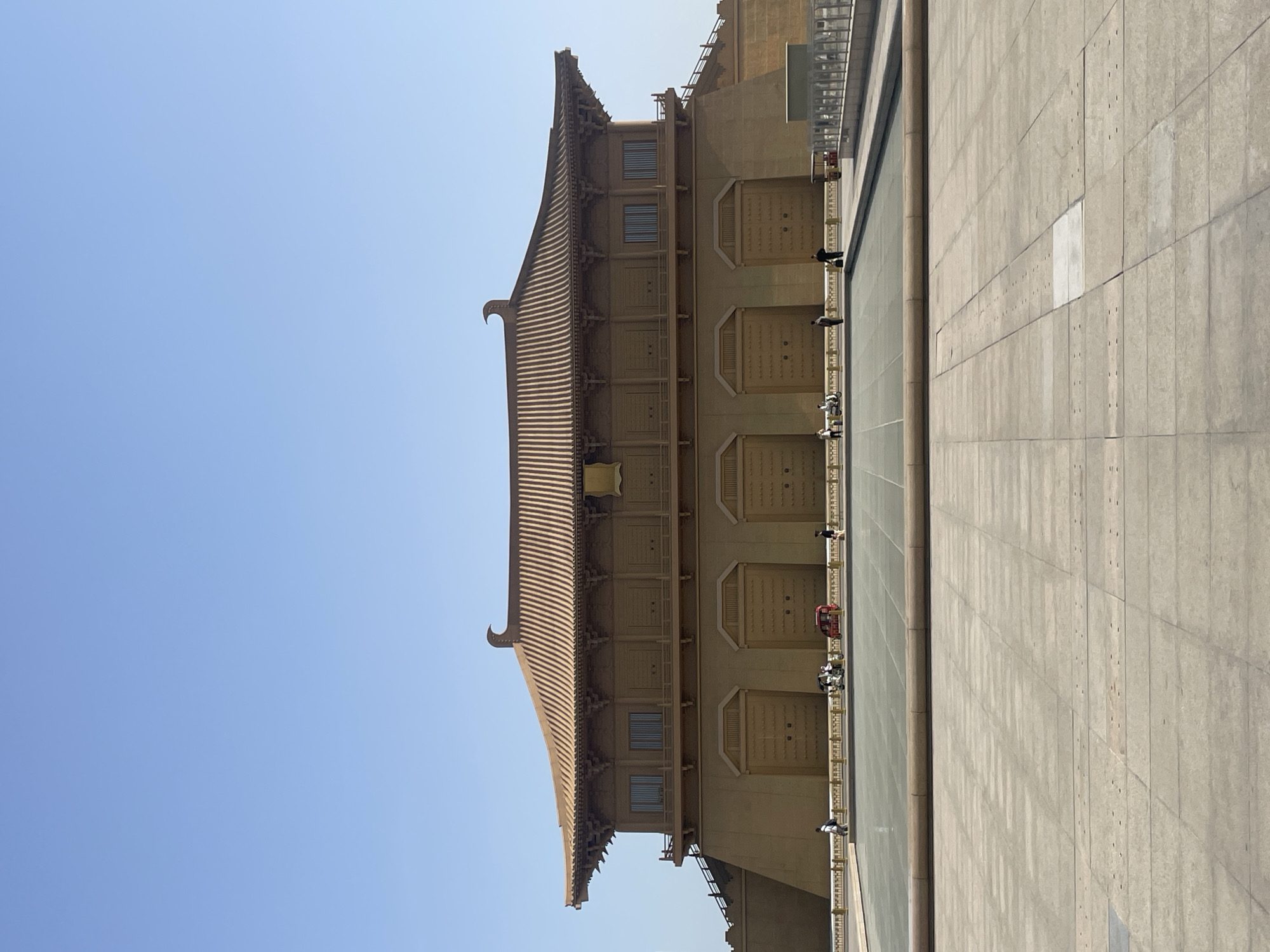 A wide, angled, outdoor shot shows a multi-storied building with a traditional curved, tiled roof, in Xincheng District, China. The building is adjacent to a raised plaza or walkway. Several people are visible near the building. The sky is clear and blue.