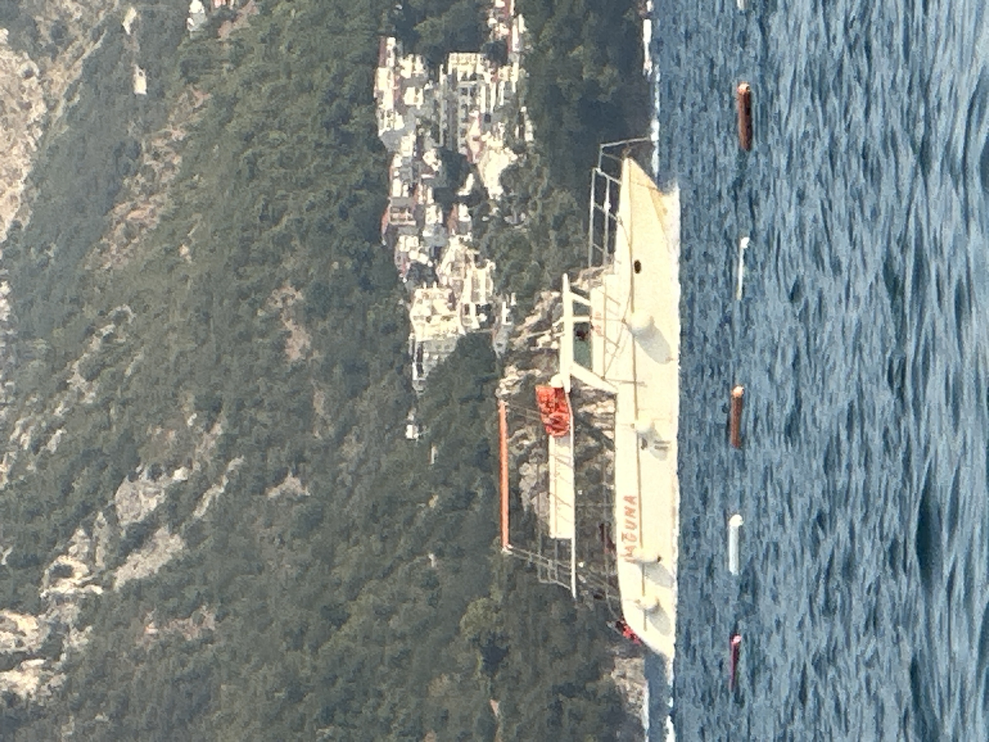 A light-colored boat, labeled "LAGUNA", is moving through the water near the city of Boreti, Montenegro. The boat has an orange canopy and several orange life preservers visible on its deck. In the background, a hillside covered in green trees rises behind a cluster of white buildings with red-tiled roofs, characteristic of the architecture in Boreti. The water in the foreground is choppy and a few floating markers are visible. The overall scene depicts a sunny day with clear visibility.
