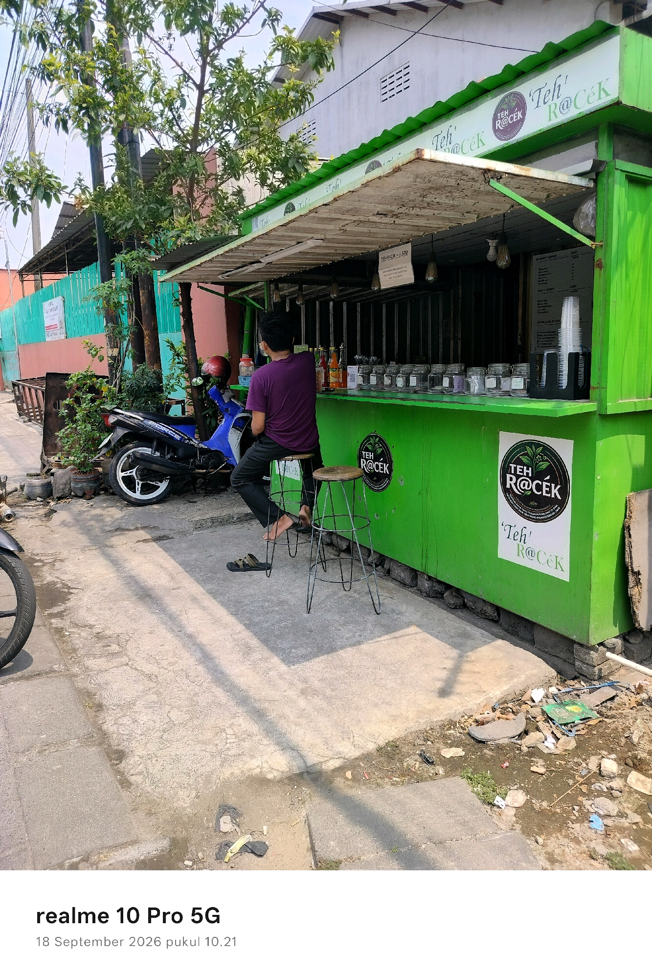 The image captures an outdoor scene in Semarang, Indonesia, featuring a bright green food stall with the prominent logo "Teh ROCEK" displayed. A person wearing a purple t-shirt and dark pants is seated on a stool in front of the stall, facing away from the camera. Beside them, a blue and black motorcycle is parked. The stall has an open counter displaying various bottled beverages and jars. A small sign is attached to the stall, indicating "TEH ROCEK = 5,000". The ground in front of the stall is paved, with scattered debris visible in some areas. The overall environment suggests a casual, urban setting typical of street-side vendors in Indonesia.