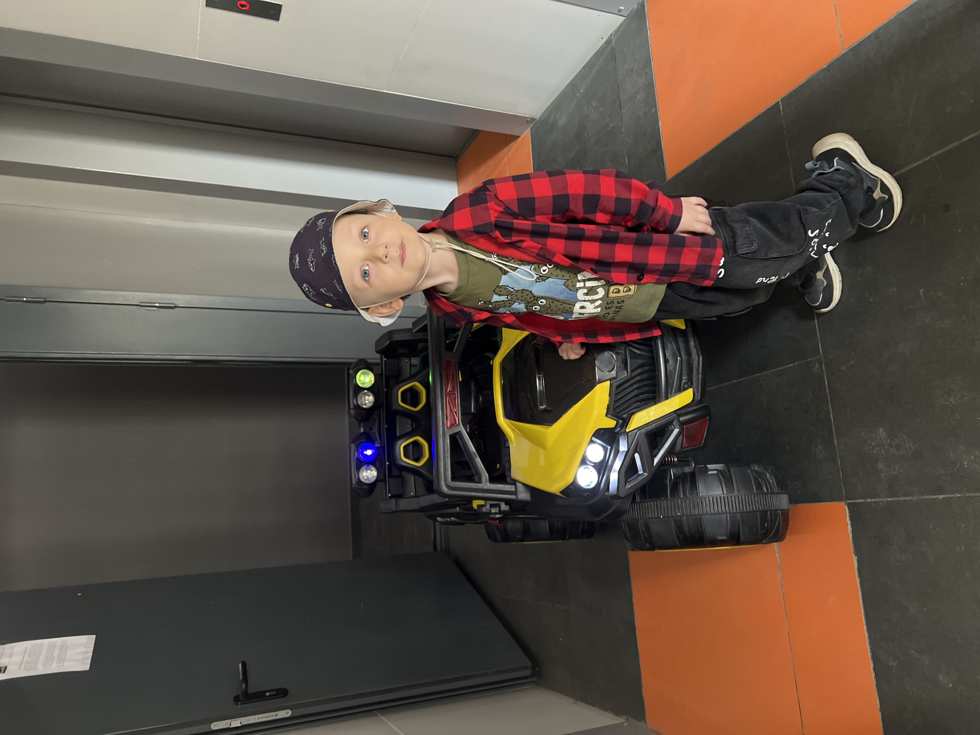 A young boy is seated in a yellow toy car in an elevator. He is wearing a printed olive-green T-shirt, a red and black plaid shirt, dark jeans, and a dark-colored hat. The toy car is equipped with illuminated headlights, and lights on the roll bars. The floor of the elevator is tiled with a pattern of black and orange squares. The image was taken in the city of Mytishchi, Russia.