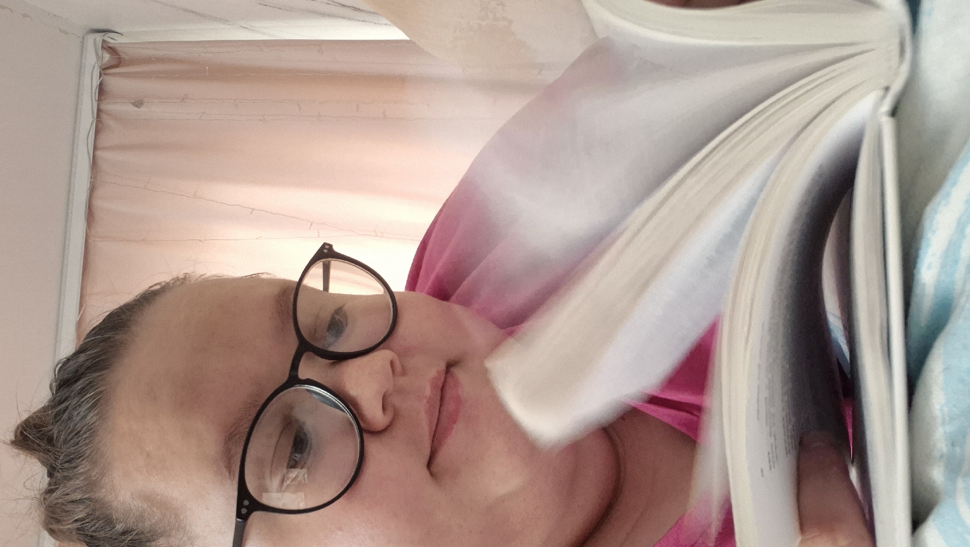 A woman with glasses is lying down, reading a book. Her head is tilted towards the camera. The background includes a peach-colored curtain or fabric and a light blue blanket. The exact location within Chaikovsky, Russia, cannot be determined from the image.