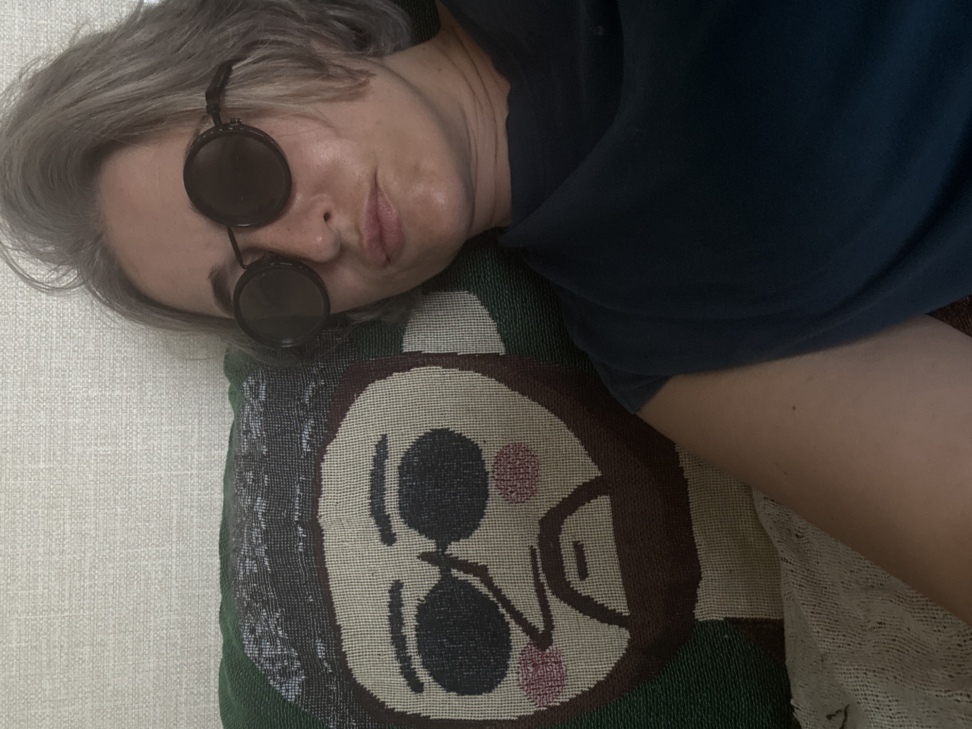 A person with short, graying hair and round, dark sunglasses is lying down, their head resting on a pillow. The pillow features a woven image of a cartoonish face with dark sunglasses, rosy cheeks, and a beard. The background appears to be a textured wall. There is no indication of the city of Kolomna, Russia in the image.