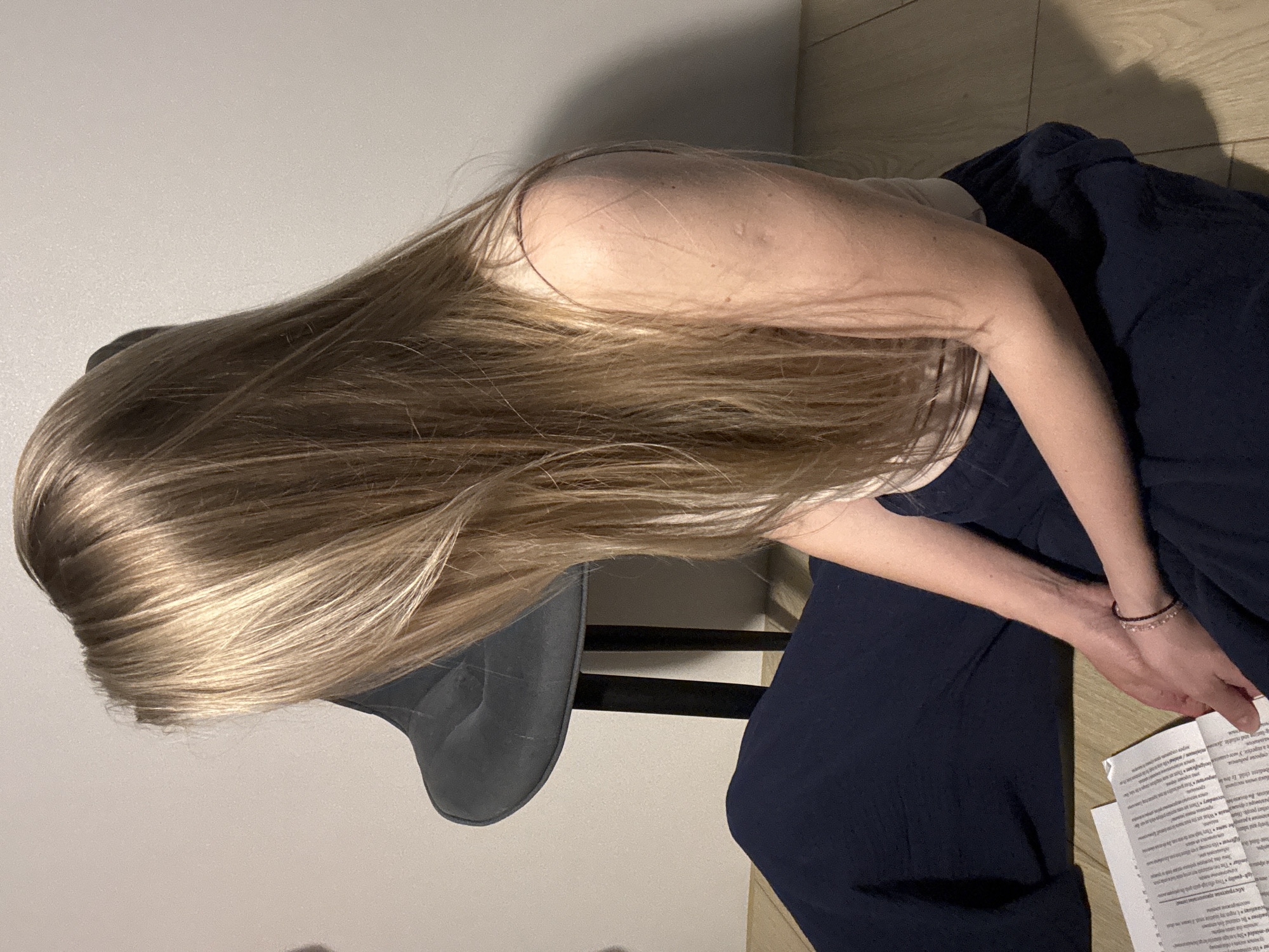 A woman with long, blonde hair is sitting on a gray chair with a black frame. She is wearing a light-colored top and dark blue pants. She is holding a piece of paper with text on it in her hands, and her arms are adorned with a beaded bracelet. The background consists of a light-colored wall and a wooden floor. The OCR indicates that some of the text on the paper is in Russian. The image does not contain any information to support the statement that the location is in the city of Chaikovsky, Russia.