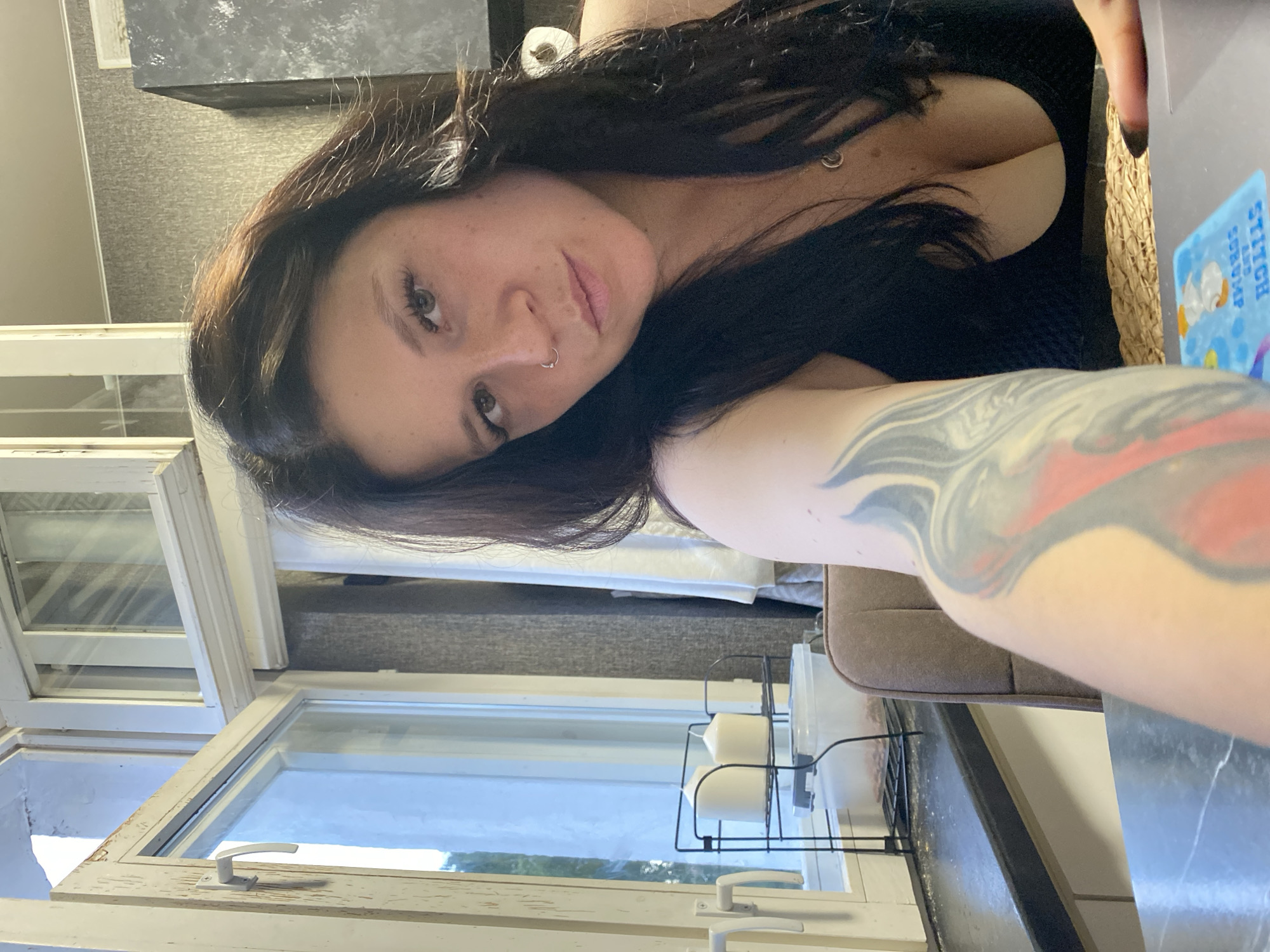 A woman with dark hair, a nose ring, and a tattoo on her arm is looking at the camera. She is wearing a black top. The scene appears to be indoors, possibly a kitchen, with a window in the background showing a glimpse of the outdoors. There are candles on a shelf by the window. The overall lighting suggests it is daytime. No specific location in Chaikovsky, Russia can be determined from the image.