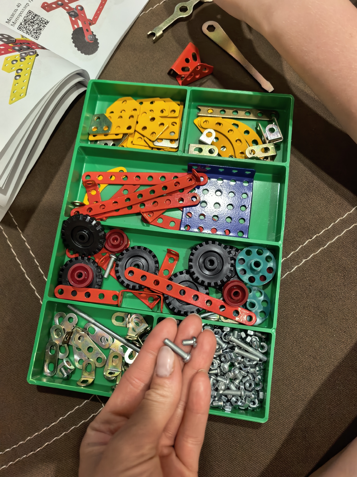 A close-up, indoor shot presents a green plastic container divided into compartments filled with construction toy parts, predominantly metal with some plastic elements, on a brown surface. A hand is holding a few screws, likely taken from the compartment containing a large pile of screws and nuts. The compartments contain various parts: yellow metal plates and curved sections, red and blue metal beams, wheels in black, red, and teal, and an assortment of metal brackets and fasteners. Tools, a wrench and a screwdriver, are placed on the surface near the container. A booklet or manual is partially visible on the left side of the image. The setting suggests a domestic environment where someone is engaging with the toy. The presence of a QR code on the booklet suggests it is a modern product. The image's location is not discernible from its contents.