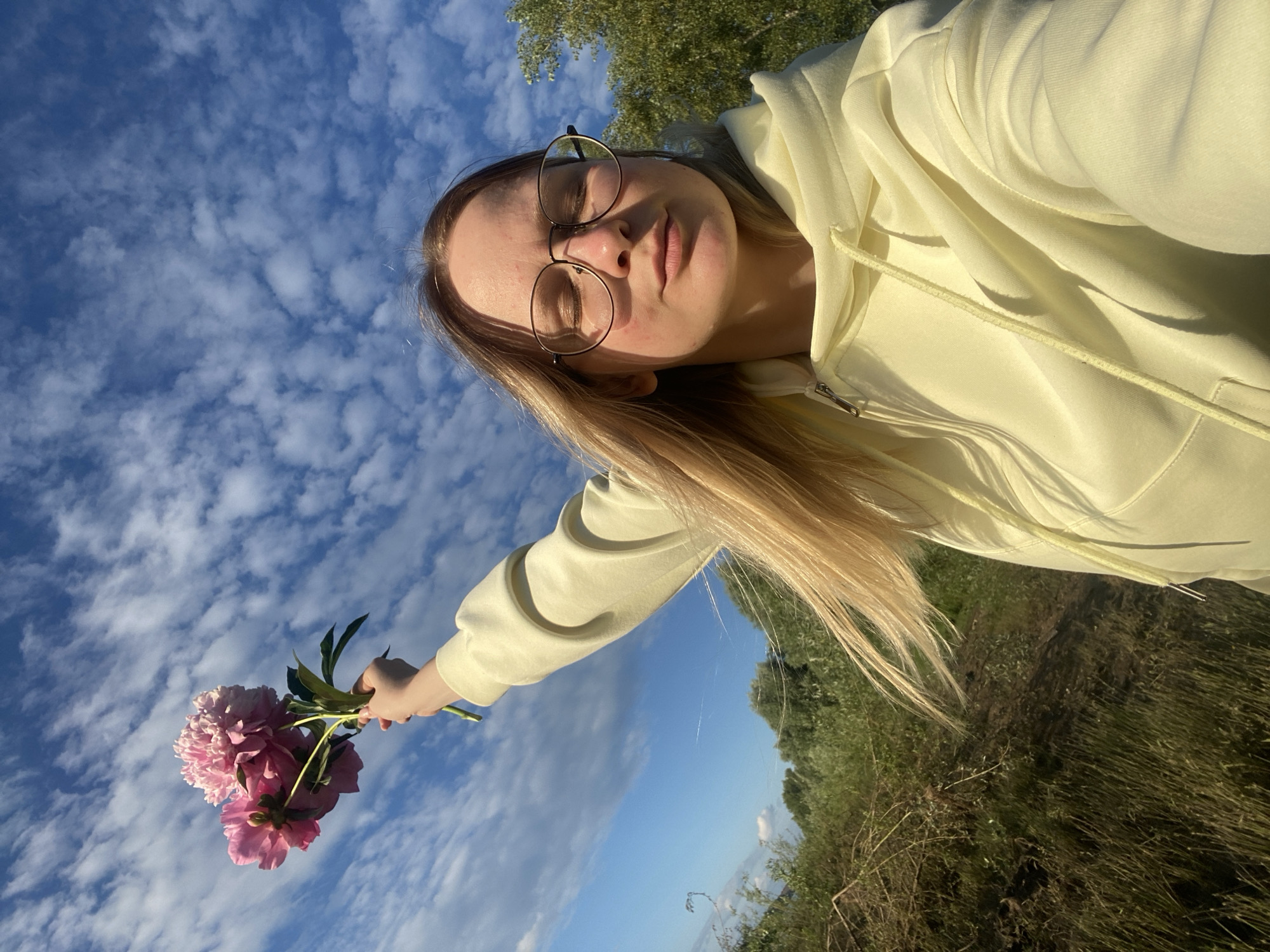 A young woman with blonde hair and glasses is taking a selfie outdoors. She is wearing a pale yellow hooded sweatshirt. In her left hand, she holds a bouquet of pink peonies. The background features a bright blue sky filled with wispy white clouds, and a line of green trees and tall grasses. The lighting suggests it is daytime, possibly late afternoon due to the warm light. The overall mood is serene and peaceful. The image appears to be taken in Chaikovsky, Russia. There is no visible text.