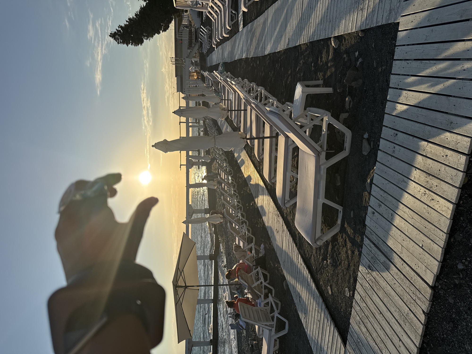The image captures a sunset scene on a pebble beach in Sochi, Russia. In the foreground, a hand is blurred as it gestures towards the sun. Rows of empty white lounge chairs and closed umbrellas are lined up on wooden pathways. In the middle ground, two women are seated on lounge chairs under a large umbrella, facing the sea. The sea is calm, with a pier extending into it in the distance. The sky is a clear blue with a few wispy clouds, and the sun is low on the horizon, casting a warm glow over the scene. A few trees are visible on the right side of the image.