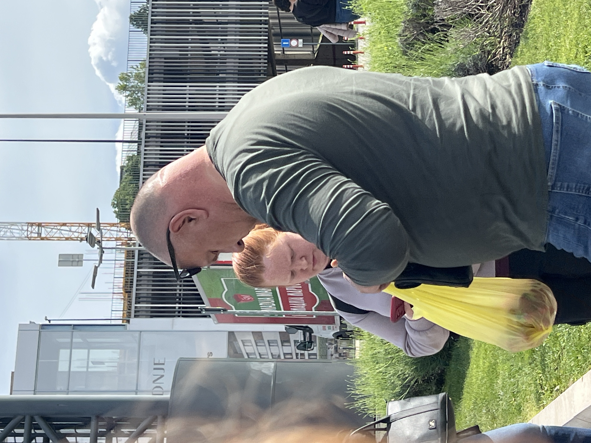 The image shows two people standing outside in Palilula Urban Municipality, Serbia. A bald man wearing sunglasses, a green long-sleeved shirt, and jeans is leaning over, looking down. Next to him is a younger person with reddish hair, wearing a light-colored sweatshirt. They are standing near a grassy area and a building with a sign in Serbian. The man is holding a yellow plastic bag containing items. A construction crane is visible in the background. A black bag is on the ground.
