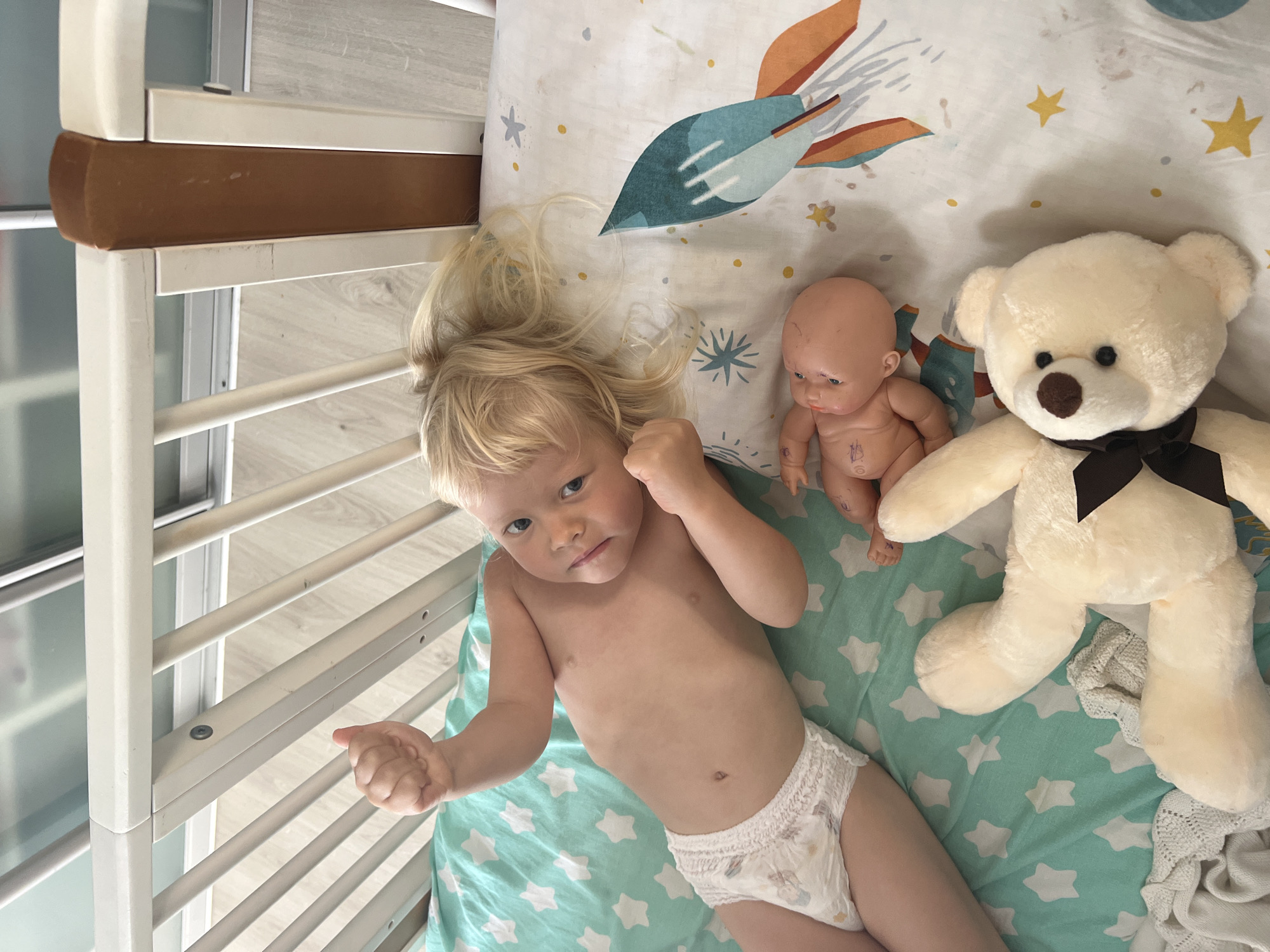 An overhead photograph captures a young, fair-skinned child with blonde hair lying on their back inside a white crib in a home in Sochi, Russia. The child, appearing to be a toddler, is wearing only a disposable pull-up diaper and looks directly at the camera with a neutral, slightly pensive expression, their fists loosely clenched.
The crib features white painted wooden slats and a brown-stained top rail. The mattress is covered by a fitted sheet displaying two distinct patterns: a light teal background with white stars beneath the child, and a white background with a large rocket ship design, yellow dots, and small stars near the child's head.
Sharing the crib space are two toys: a small, bald, light-skinned doll with faded markings on its torso, and a cream-colored teddy bear with a brown snout and a dark brown bow around its neck. A light-colored, possibly knitted, blanket is bunched near the teddy bear's feet.
The background outside the crib reveals light-colored flooring, likely wood laminate, and part of a modern wardrobe or sliding glass doors with a frosted finish. The bright, even lighting suggests it is daytime indoors. The scene depicts the child resting or playing quietly with their toys in the crib. No legible text is visible in the image.