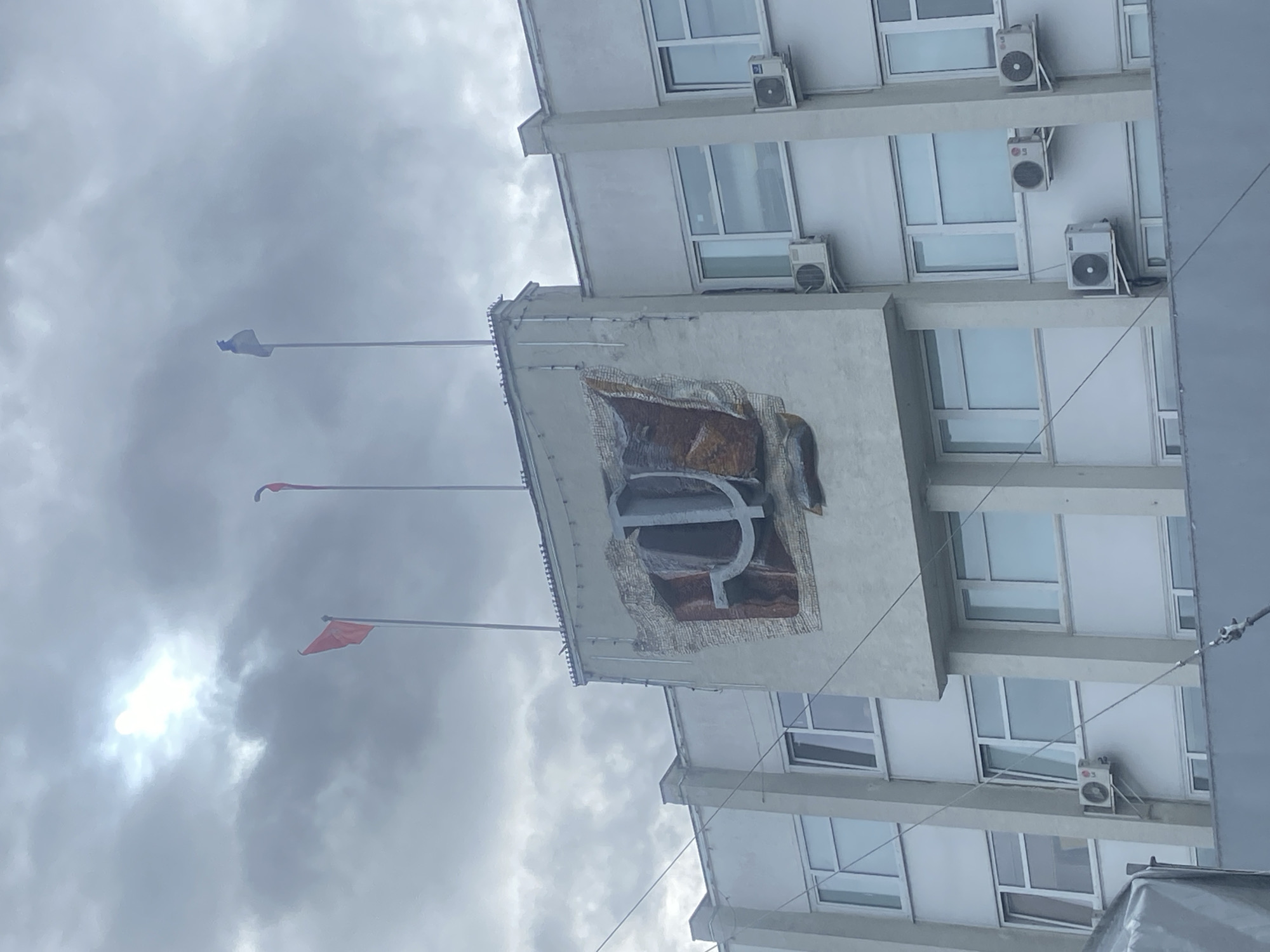 The image shows a multi-story building with a large emblem on an extended section. The emblem appears to be a stylized letter "P" or Cyrillic "R", possibly with mosaic or relief details. Three flagpoles with flags are visible above the emblem, extending into a cloudy sky. The building has multiple windows and several air conditioning units. The location is in Sergiyev Posad, Russia, as indicated by the emblem that symbolizes the city.
