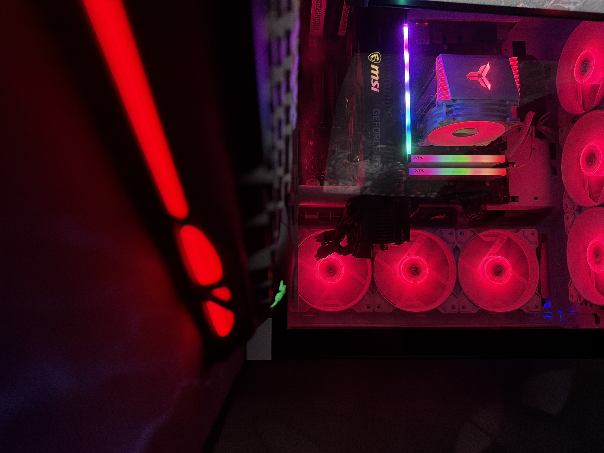 The image is a close-up, dimly lit photograph of the interior of a gaming computer. The dominant color scheme is red, emitted by the computer's internal lighting, which includes fans and LED strips. In the foreground, a blurred keyboard is visible, with some of its keys reflecting red light. The background features components of the computer, including a large CPU cooler with a stylized logo, RAM sticks with RGB lighting, and a graphics card with "MSI GEFORCE RTX" branding illuminated. Several large fans, likely case fans, are visible on the right side of the computer, all glowing red. The overall impression is of a high-performance gaming setup with extensive RGB customization. There are no people, specific city locations, or discernible environmental context beyond the computer itself.