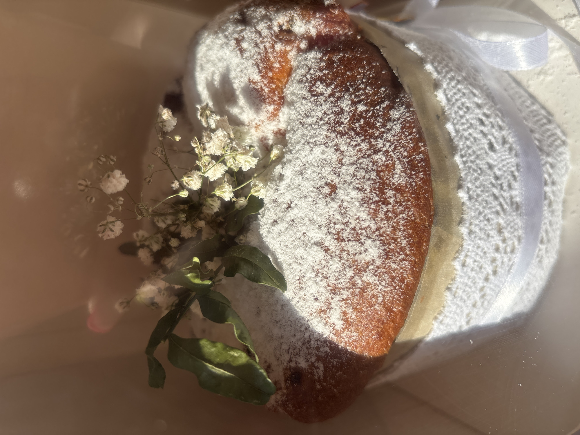 A close-up shot shows a loaf of bread heavily dusted with powdered sugar, wrapped in what appears to be a lacy fabric and tied with a white ribbon. A small arrangement of baby's breath and greenery decorates the left side of the bread. The bread and decorations are situated inside a light-colored container. The lighting suggests the photo was taken indoors with natural light. There is no indication of the location being Русский Вожой, Russia.
