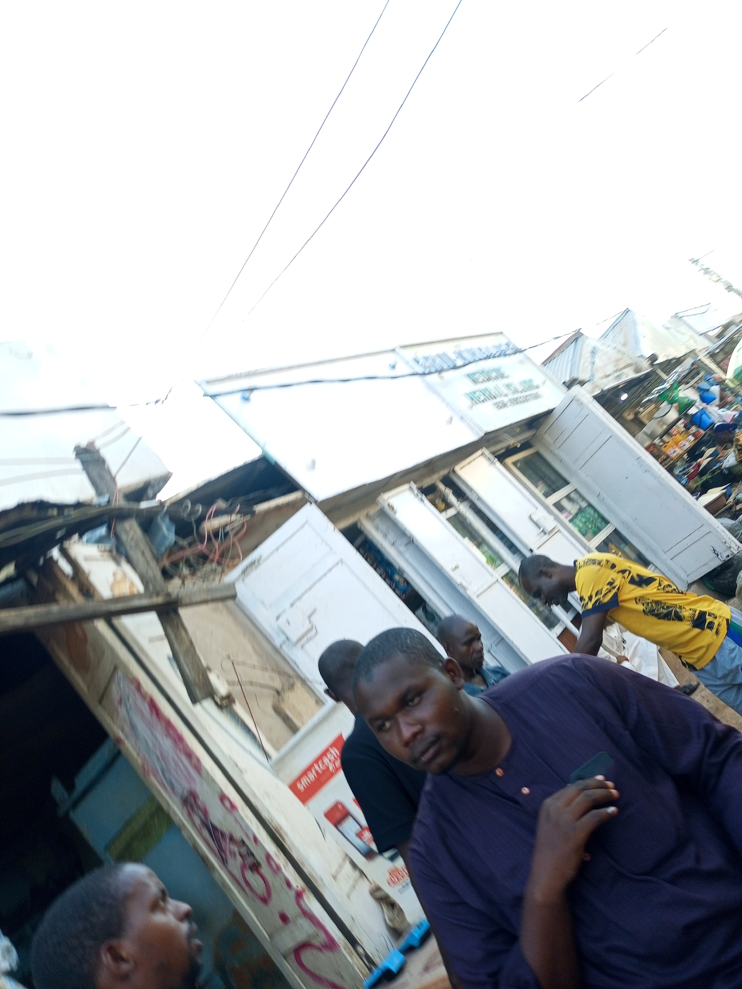 The image depicts a street scene in Tempure, Nigeria, with a focus on a damaged building or structure. Multiple people are visible, some appearing to be interacting with the building or its surroundings. One man in the foreground wears a dark purple garment and is looking towards the camera. Another man in a yellow shirt is seen in the middle ground, seemingly engaged in some activity. The building itself shows signs of damage, with debris and exposed wiring. Signs on the building are partially visible, one appearing to read "Smartcash". The overall impression is one of a bustling but potentially disorganized or recovering urban environment. The image is taken from a low angle, looking upwards towards the sky and the damaged structure.