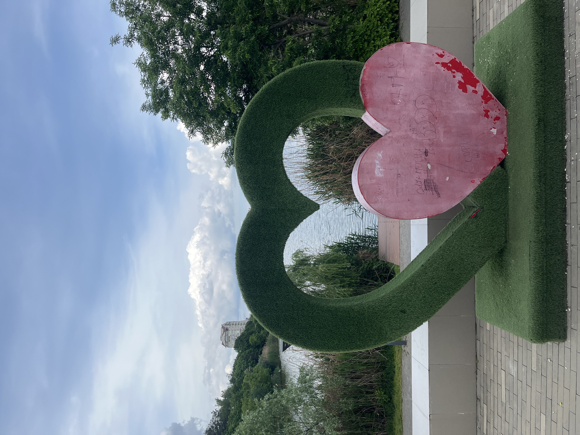 A green heart-shaped arch with a smaller red heart attached is located in a park in Krasnodar, Russia. The arch is covered in artificial turf. The red heart is worn and has graffiti on it. The park has trees and is next to a body of water. A building can be seen in the distance. The sky is partly cloudy. A paved path is visible in the foreground.
