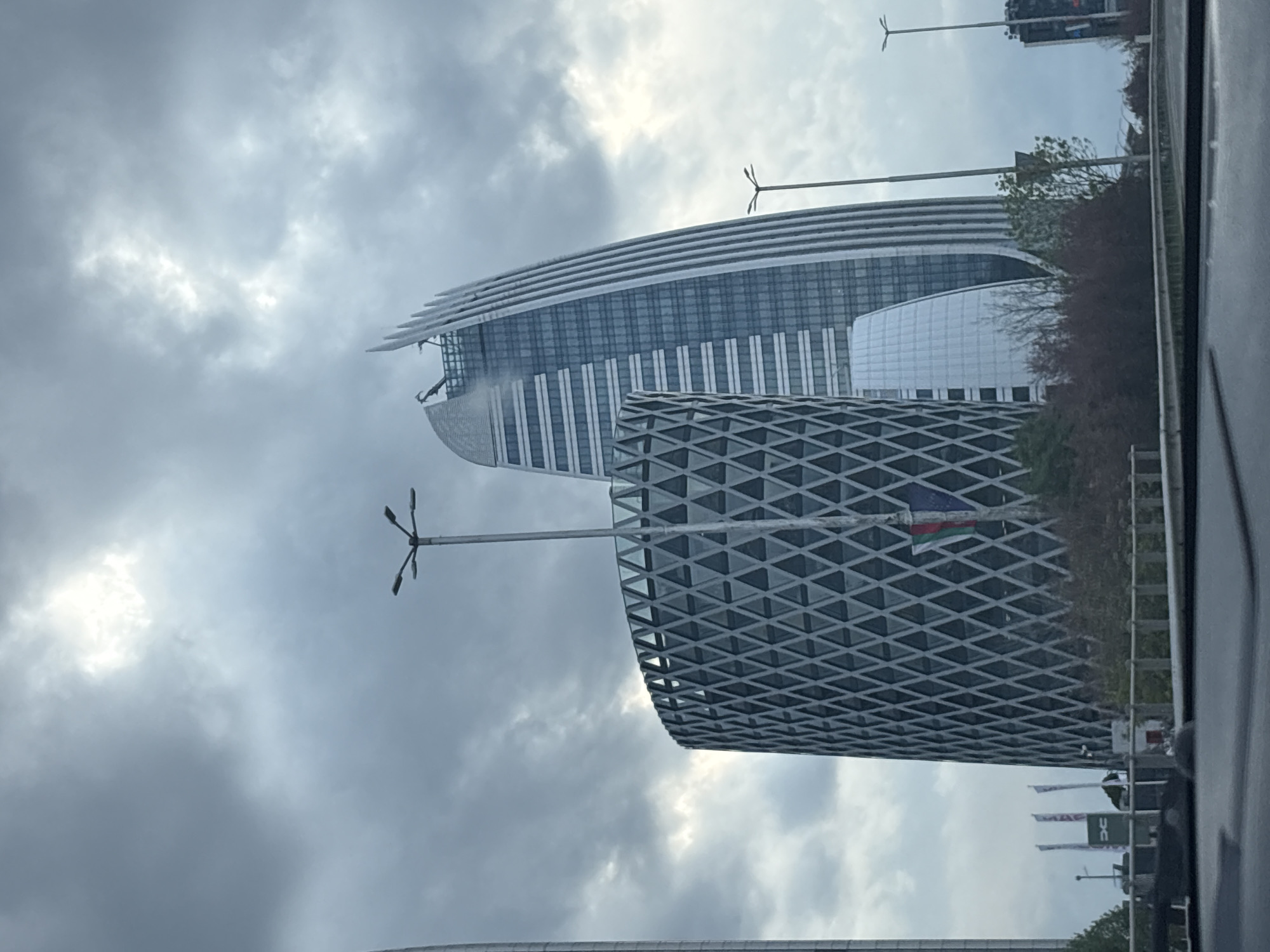 The image, taken from within a vehicle, presents a modern cityscape under an overcast sky, with the perspective rotated 90 degrees to show buildings vertically.
The scene is dominated by several contemporary buildings. In the midground, a distinctive skyscraper features a curved, layered roofline composed of multiple horizontal fins or slats, giving it an elegant, sweeping silhouette. Its main facade consists of expansive glass panels punctuated by vertical white elements. Adjacent to this, a smaller, rounded white structure with a grid pattern is partially visible.
In the foreground, another large building captures attention with its intricate geometric facade, formed by a repeating diamond-shaped grid. A flag, likely the national flag of Bulgaria (displaying white, green, and red stripes), is attached to this building's exterior, visible on the left side of its textured surface.
The sky is heavily clouded, indicating an overcast daytime, though patches of brighter light suggest the sun is attempting to break through. Tall streetlights with multi-pronged fixtures are visible throughout the frame. Sparse trees and vegetation line what appears to be a road or urban pathway at the base of the structures. The dark frame of a car window is visible on the right edge of the image, reinforcing the in-vehicle perspective. Further down, several flags bearing the text "DC" are also present. The scene depicts a modern urban environment in Sofia, Bulgaria.